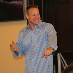 A smiling man in a blue checkered shirt standing in front of a dark background, gesturing with his hands.