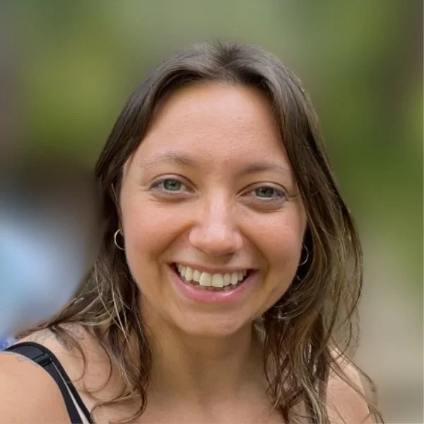 A young woman smiling outdoors with a blurred green background.