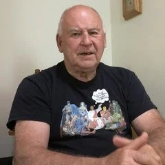 Older man with short white hair sitting indoors, wearing a black t-shirt with a colorful graphic on the front.