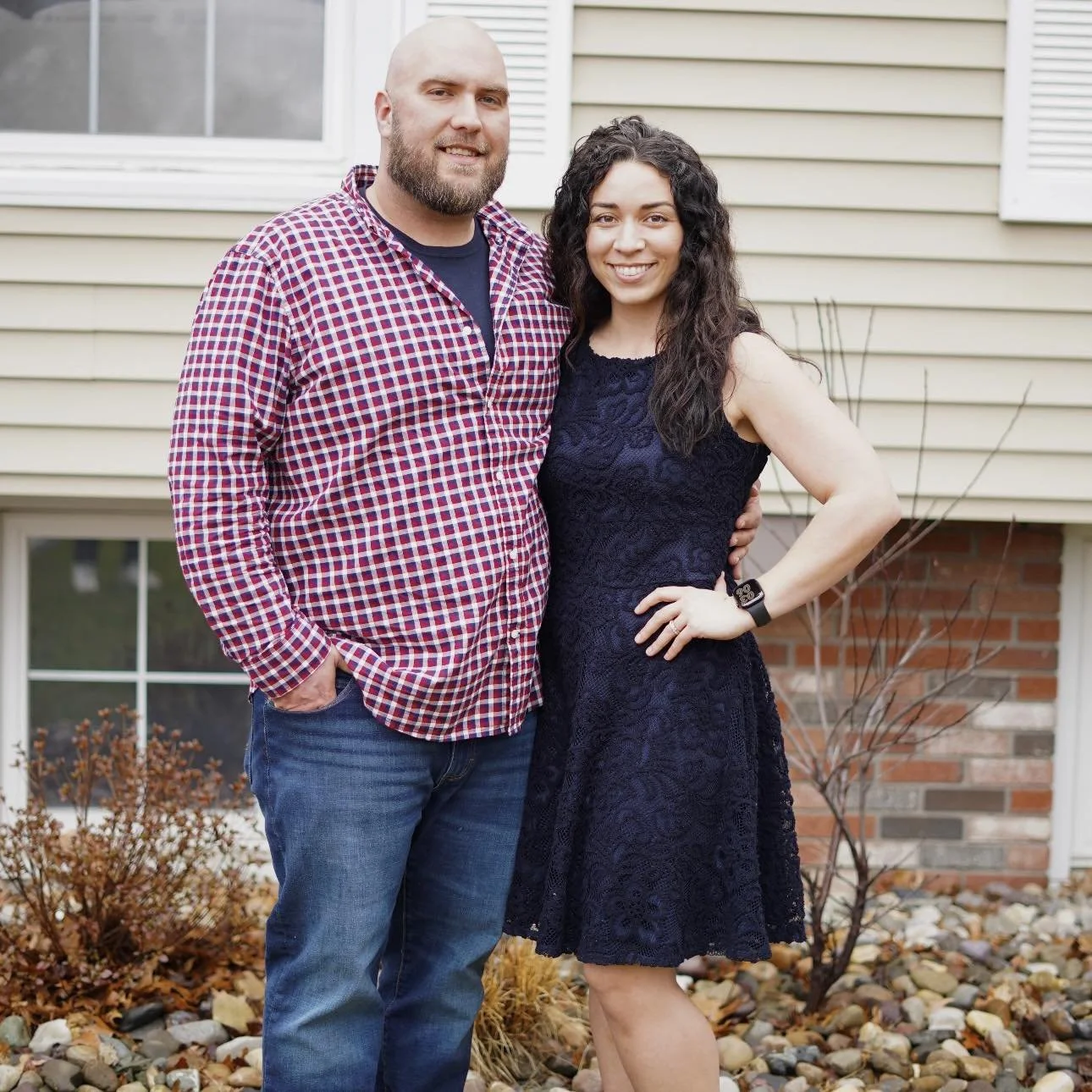 A happy couple standing outside a house with beige siding and a brick foundation. The man has a beard, is bald, and wearing a red checkered shirt and jeans. The woman has curly dark hair, wearing a black lace dress and a fitness tracker on her wrist. They are smiling and standing close together.