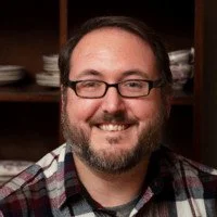 A man with glasses and a beard smiling in front of a bookshelf with plates and bowls.