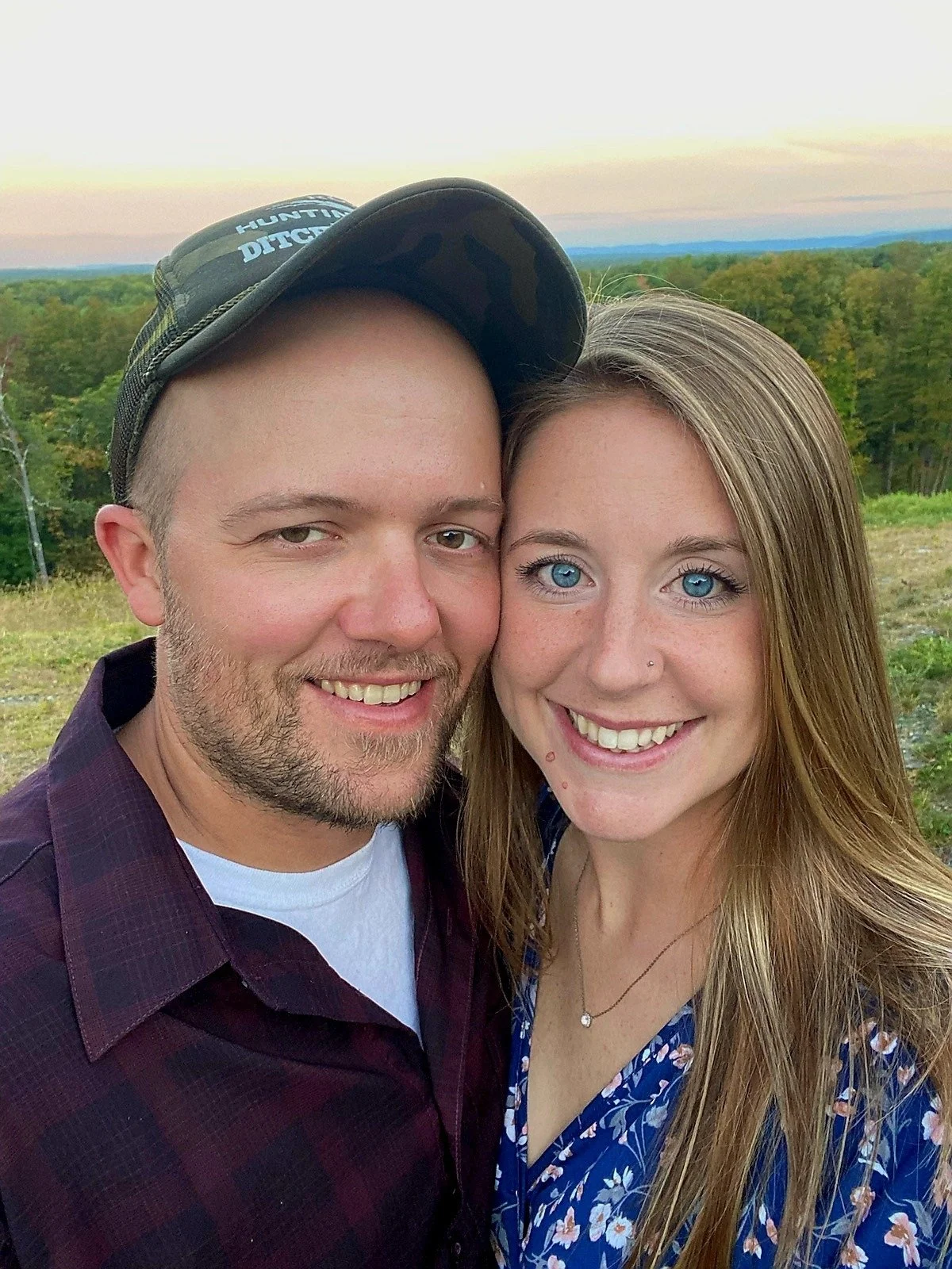 A smiling couple taking a selfie outdoors in a scenic landscape with green trees and a pastel-colored sky.