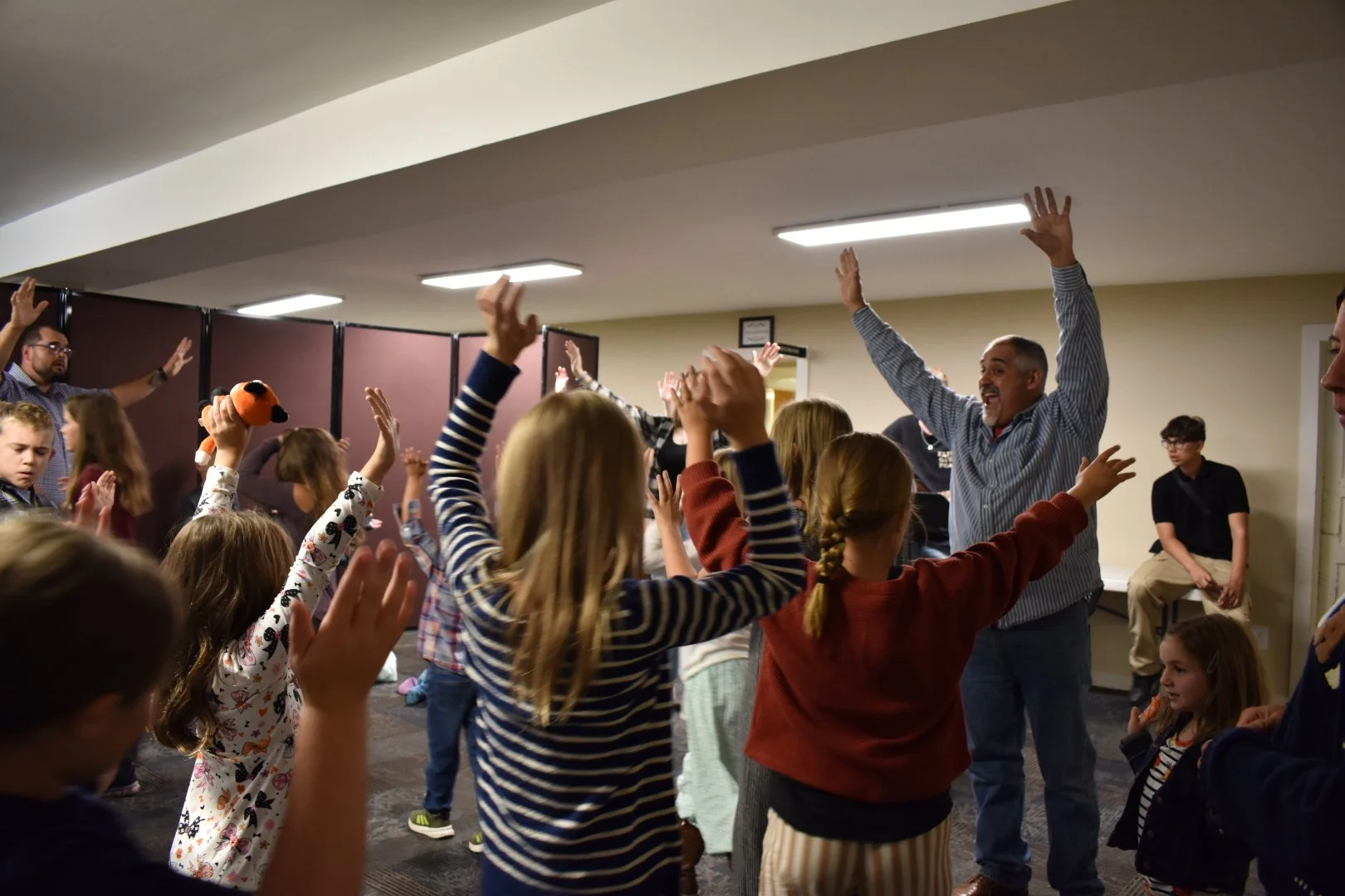 Group of children and adults in a room raising their hands, participating in an activity or game, under ceiling lights.