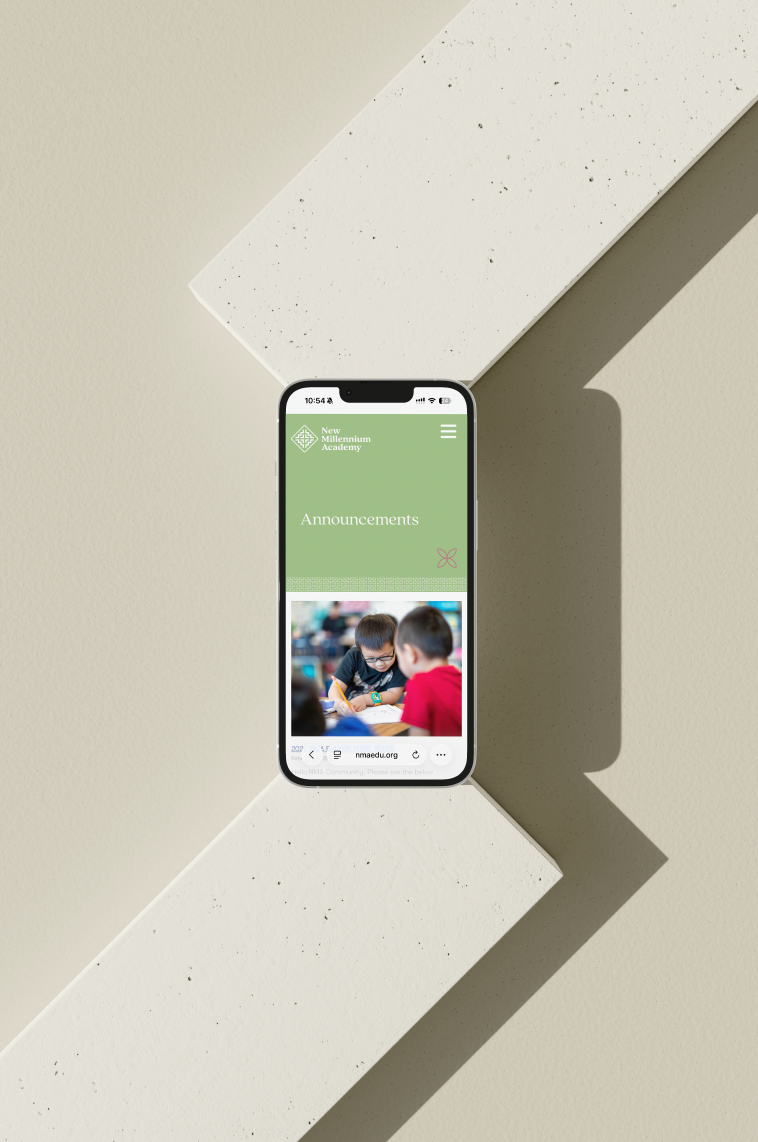 A smartphone displaying the New Millennium Academy website with an announcement page and a photo of two children engaging in learning activities, set on a beige background with white textured surfaces.