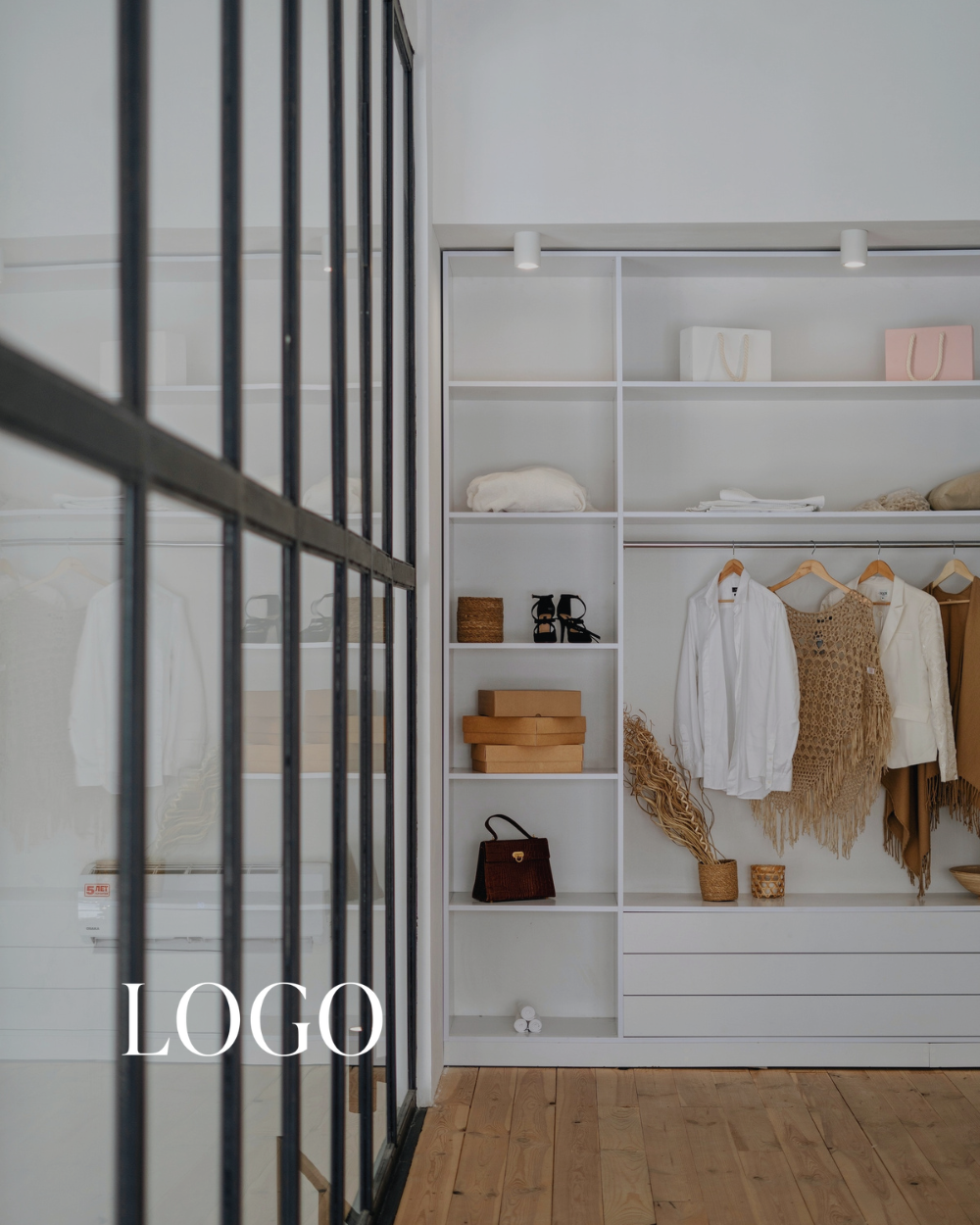 Empty white closet with hanging clothes, shoes, bags, and shelves, in a room with wooden flooring and white walls. A black metal-framed glass door is on the left. The closet contains whites, beige, and brown items, with some decorative pieces and storage boxes.