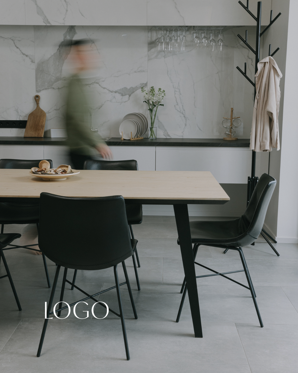 Modern dining room with a wooden table and black chairs, a marble wall with kitchen accessories, and a coat rack with a beige coat hanging.