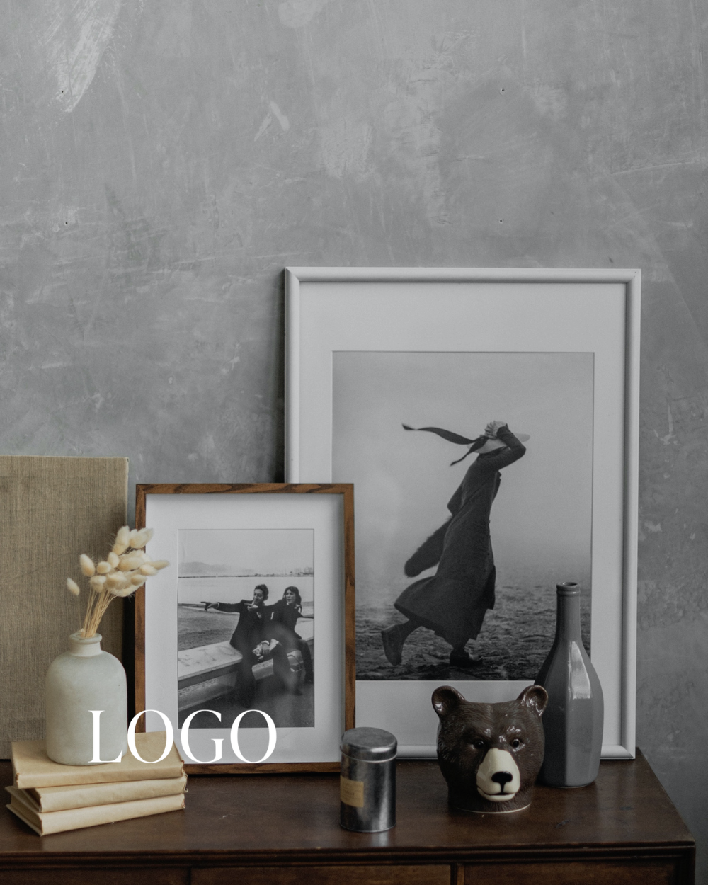 Black and white photographs in frames on a wooden table with decorative items including a white vase with dried flowers, a bear-shaped container, a small black candle, and two vases, set against a textured gray wall.