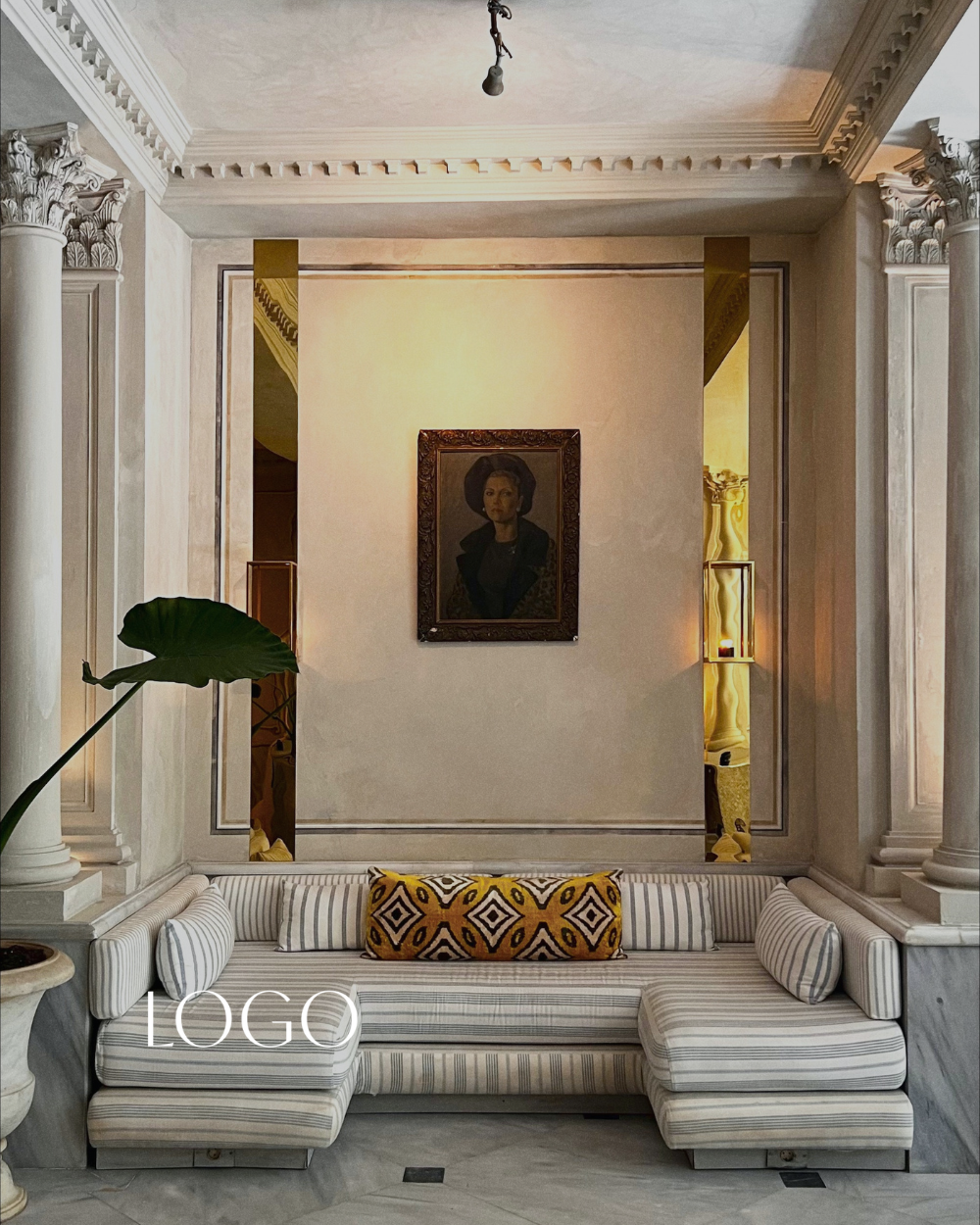 Interior of a grand room with classical architecture featuring ornate columns and decorative moldings. A striped sofa with a yellow patterned pillow and cylindrical cushions is positioned in front of a wall with a portrait of a woman. A large green leaf is visible on the left, and there are two tall, narrow wall sconces with warm lighting on either side of the portrait.