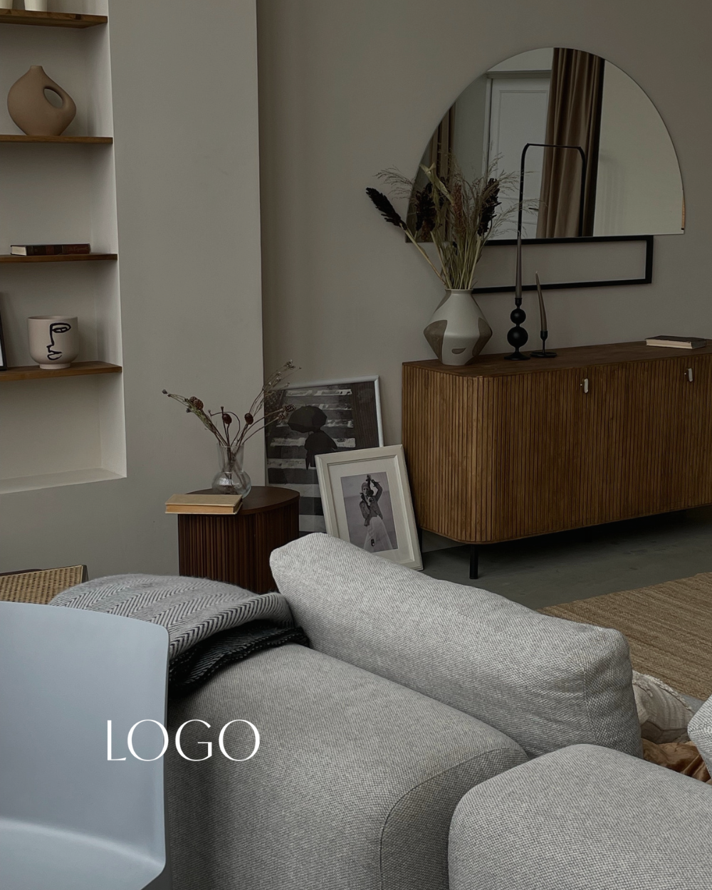A modern living room corner with a beige sofa, a wooden side table with a glass vase and books, a wooden cabinet with decorative objects and framed artwork leaning against it, a large round mirror with a semi-circular top behind the cabinet, and a minimalist aesthetic with neutral tones and decorative vases on display.