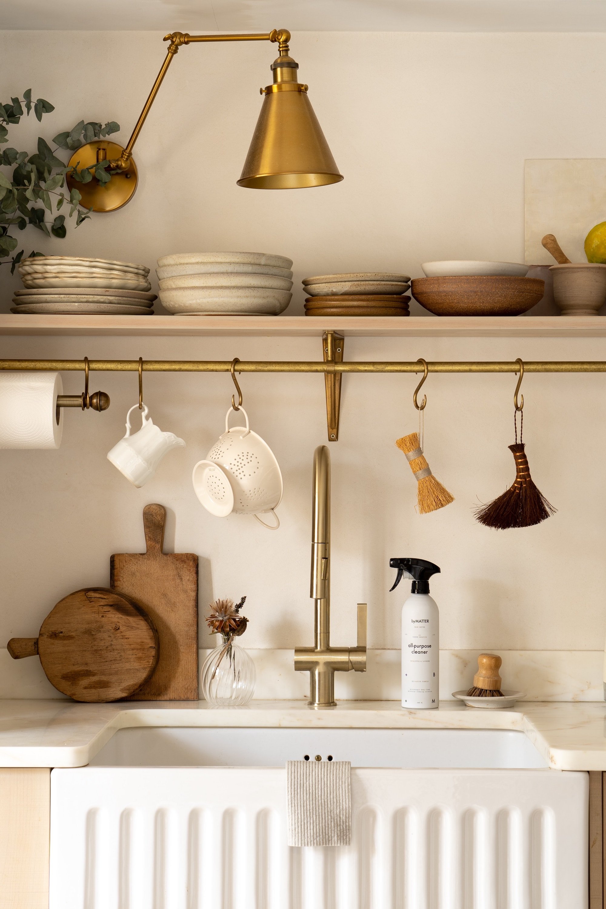 Kitchen sink with a gold faucet, white backsplash, wooden cutting boards, a vase with dried flowers, a bottle of all-purpose cleaner, hanging cups, a towel on the sink, and a brass wall-mounted lamp with a gold shade.