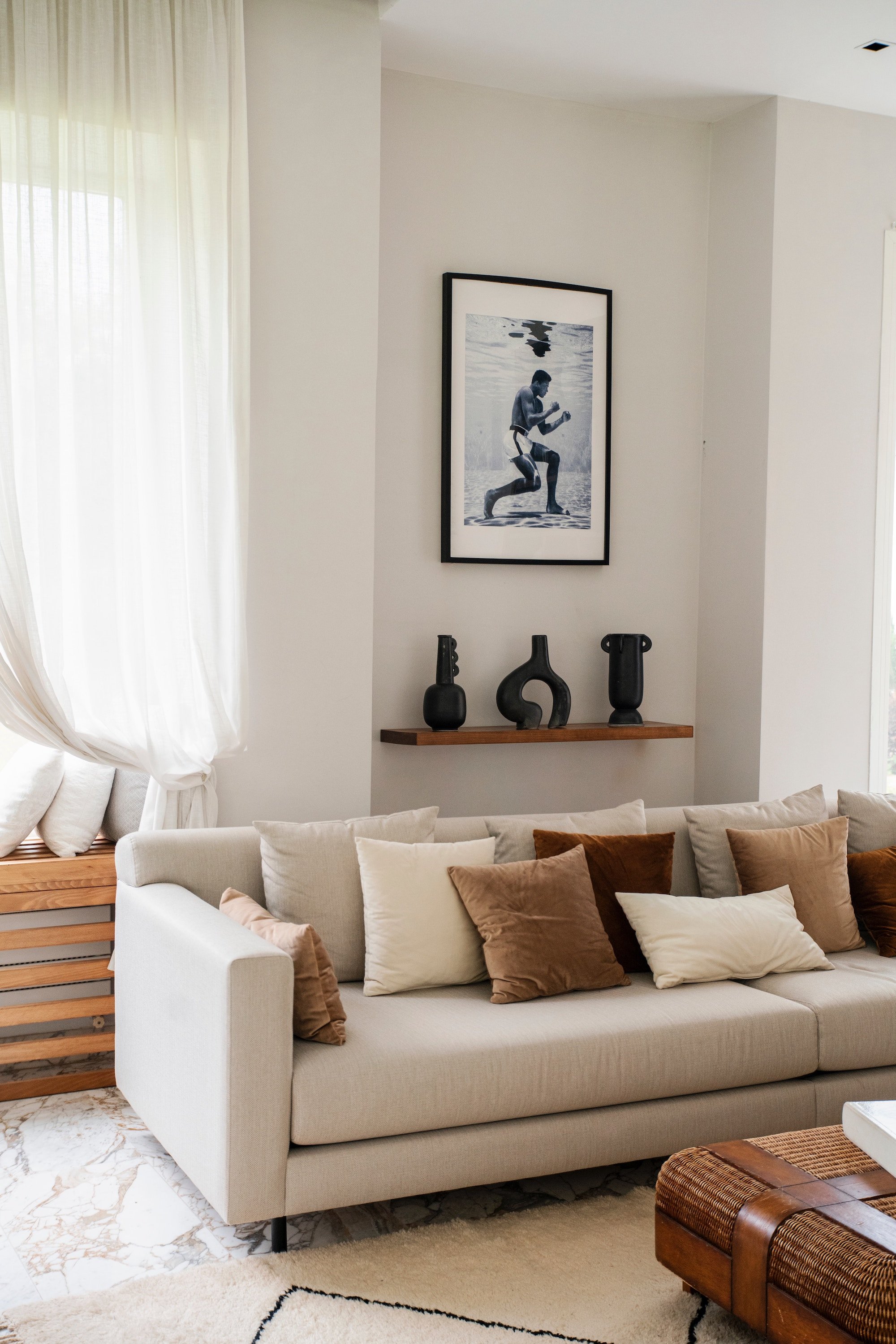 A beige sofa with multiple brown and beige pillows in a living room. A wooden table with decorative vases hangs above it. A framed picture of a person boxing underwater is on the wall. Part of a window with sheer curtains is visible on the left.