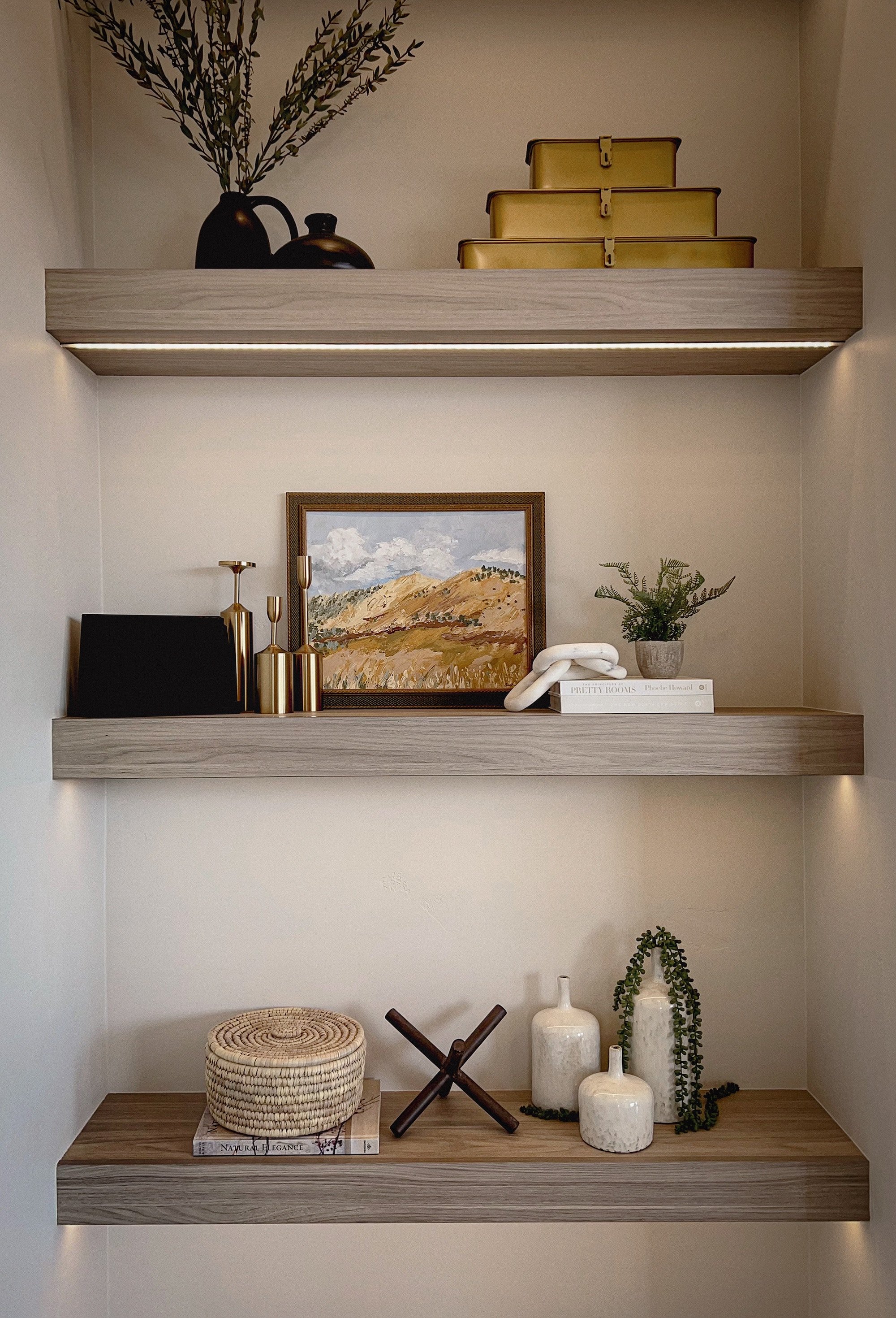 Three floating shelves with decorative items on a beige wall. The top shelf has a black vase with branches and three gold-colored boxes. The middle shelf has gold candle holders, a landscape painting, and a small potted plant. The bottom shelf has a woven basket, a book titled 'Natural Elegance,' a dark brown chair, and three white vases with greenery.