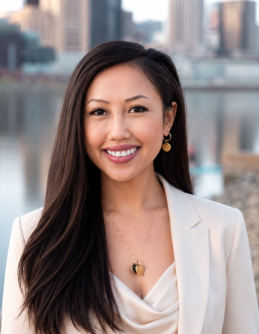 A smiling woman with long dark hair wearing a cream blazer, gold jewelry, and standing outdoors in front of a cityscape with buildings and water.