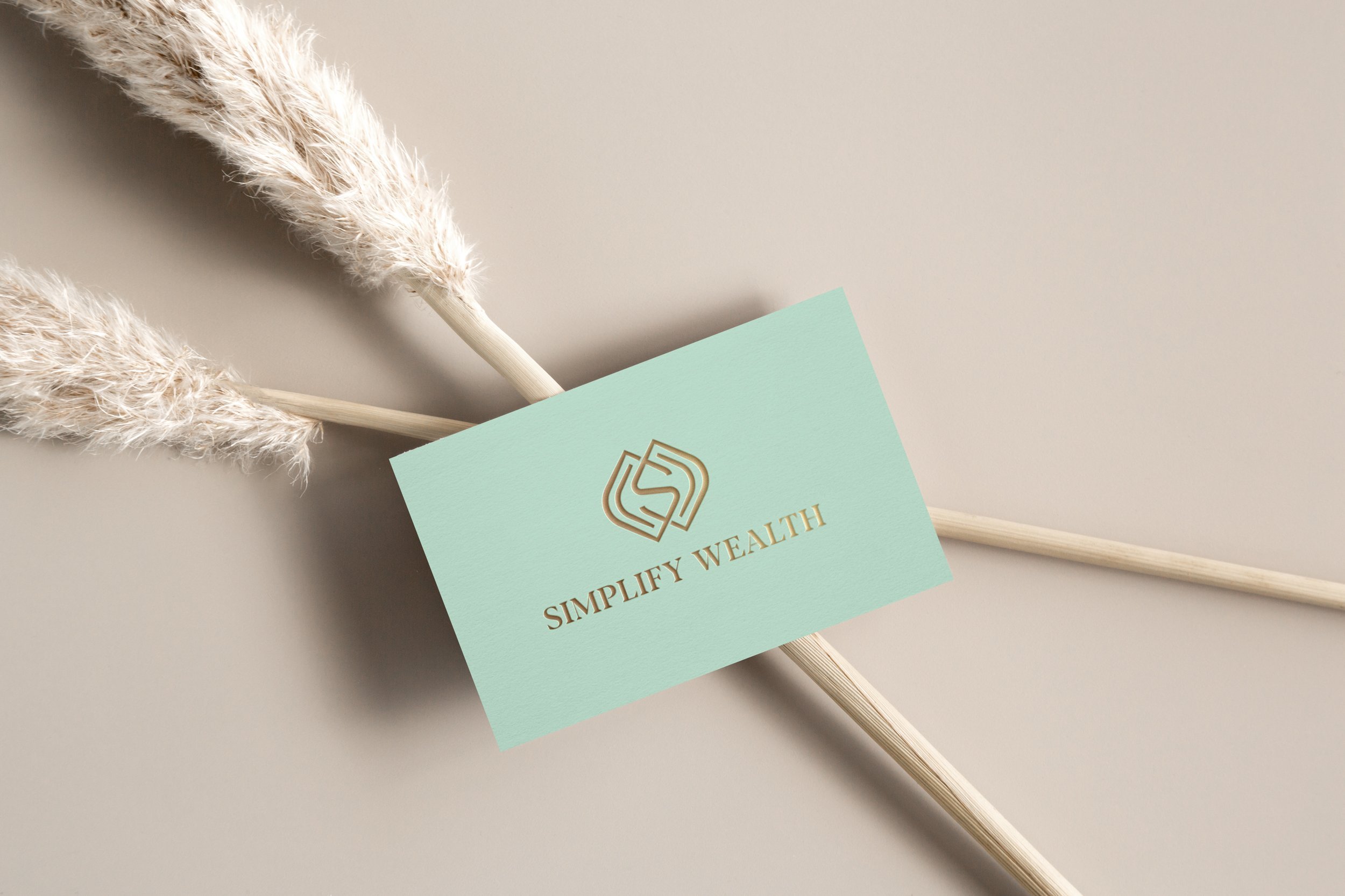 Two fluffy, beige pampas grass stems crossing each other on a beige background, with a light green business card that reads 'SIMPLIFY WEALTH' partially tucked underneath the grass.