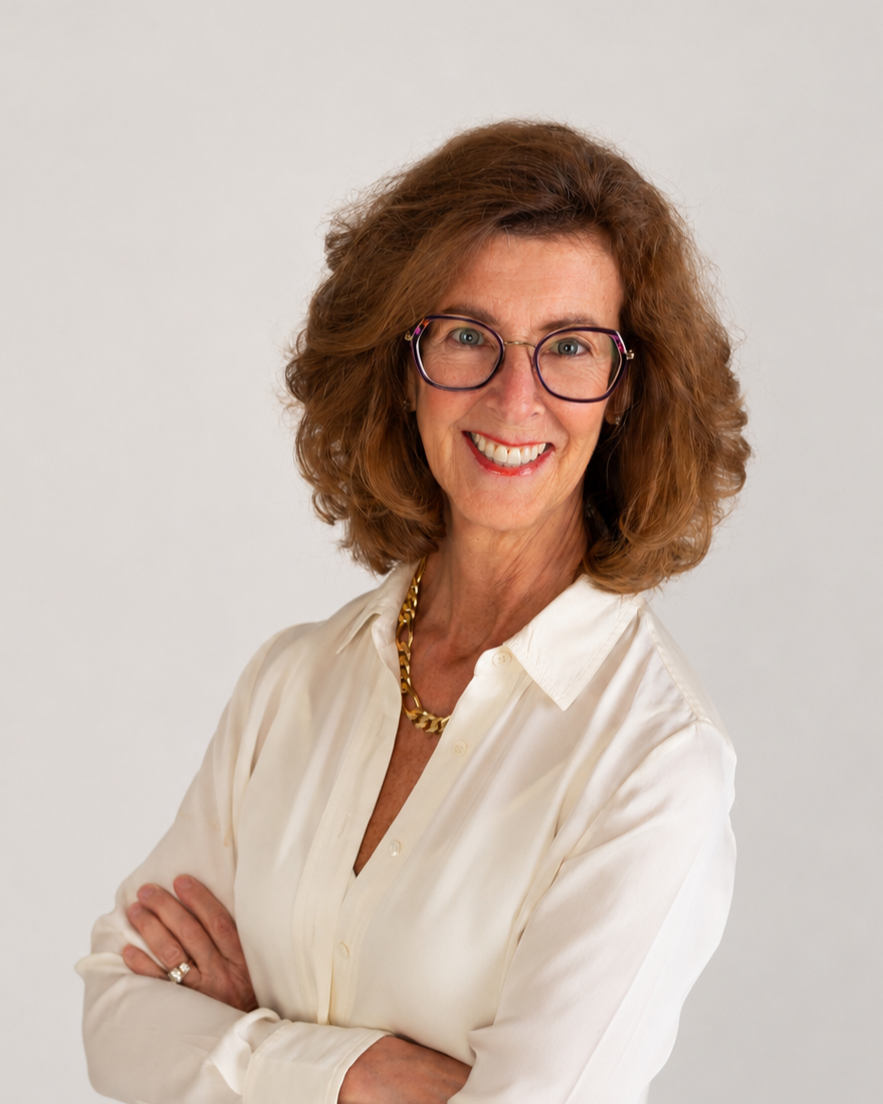 A middle-aged woman with curly, shoulder-length auburn hair and glasses, smiling with arms crossed, wearing a cream-colored blouse and a gold chain necklace.