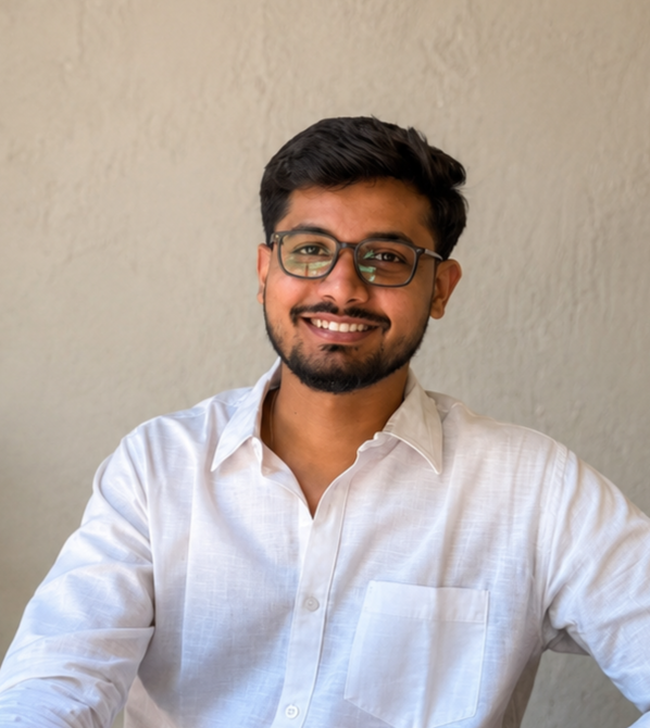A young man with dark hair and glasses, smiling, wearing a white shirt, sitting against a beige wall.