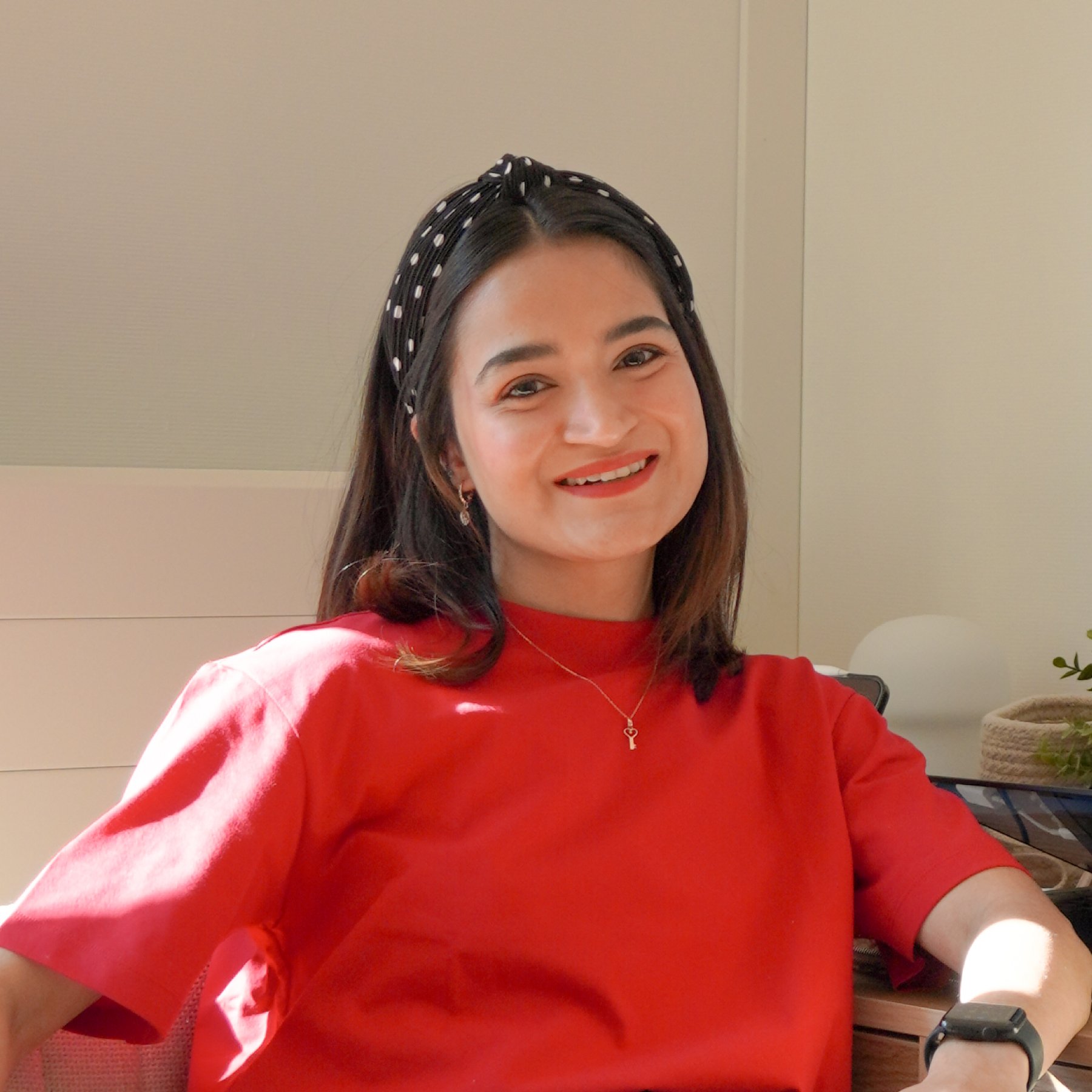 A woman smiling while sitting at a desk, wearing a red top and a black headband with white polka dots. She has a necklace with a key pendant and is wearing a smartwatch on her left wrist. There are plants and office items in the background.