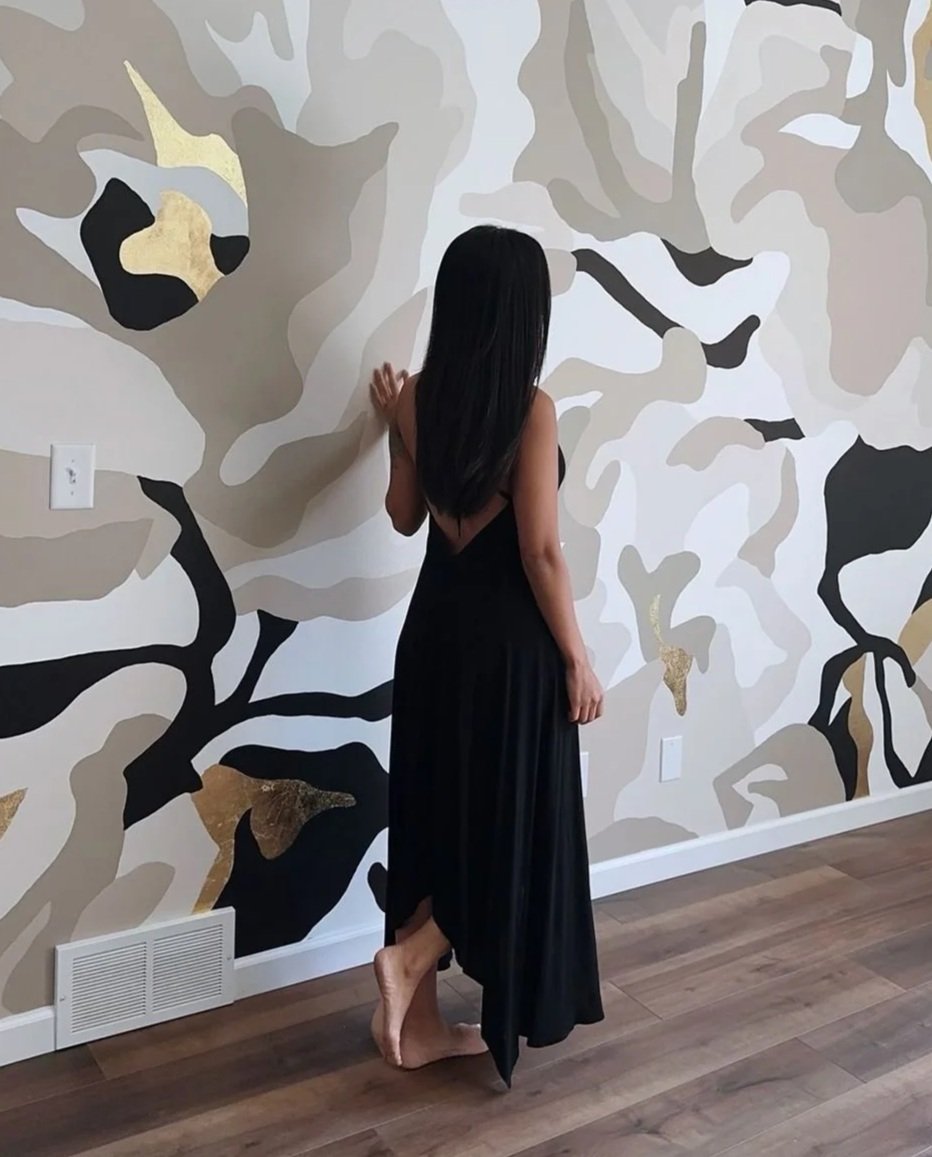 A woman with long black hair, wearing a black dress, is standing barefoot facing a large abstract floral mural on a wall. The mural features swirling patterns in shades of beige, white, black, and gold.