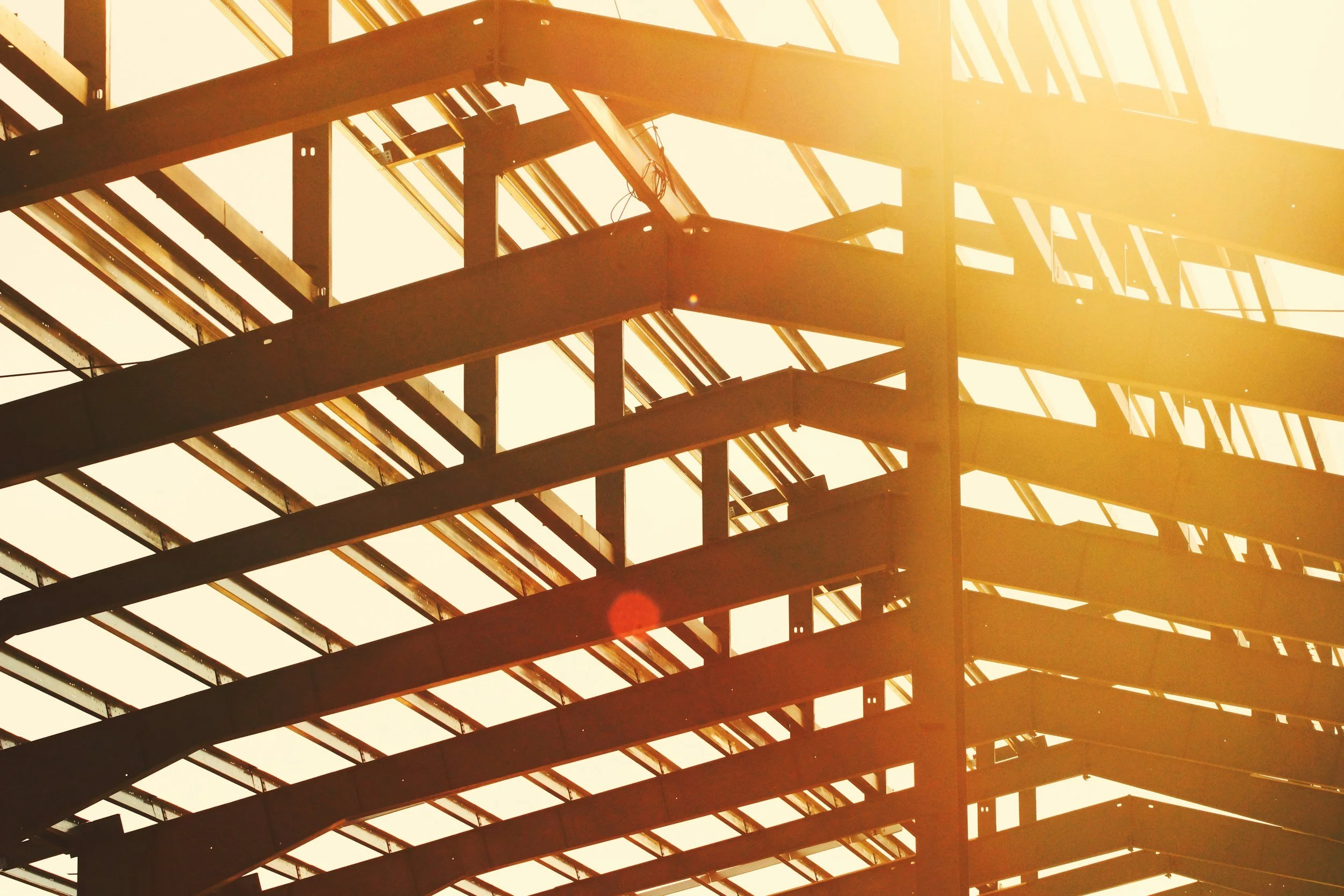 Looking up at the framed structure of a building with steel beams and glass panels, illuminated by sunlight.