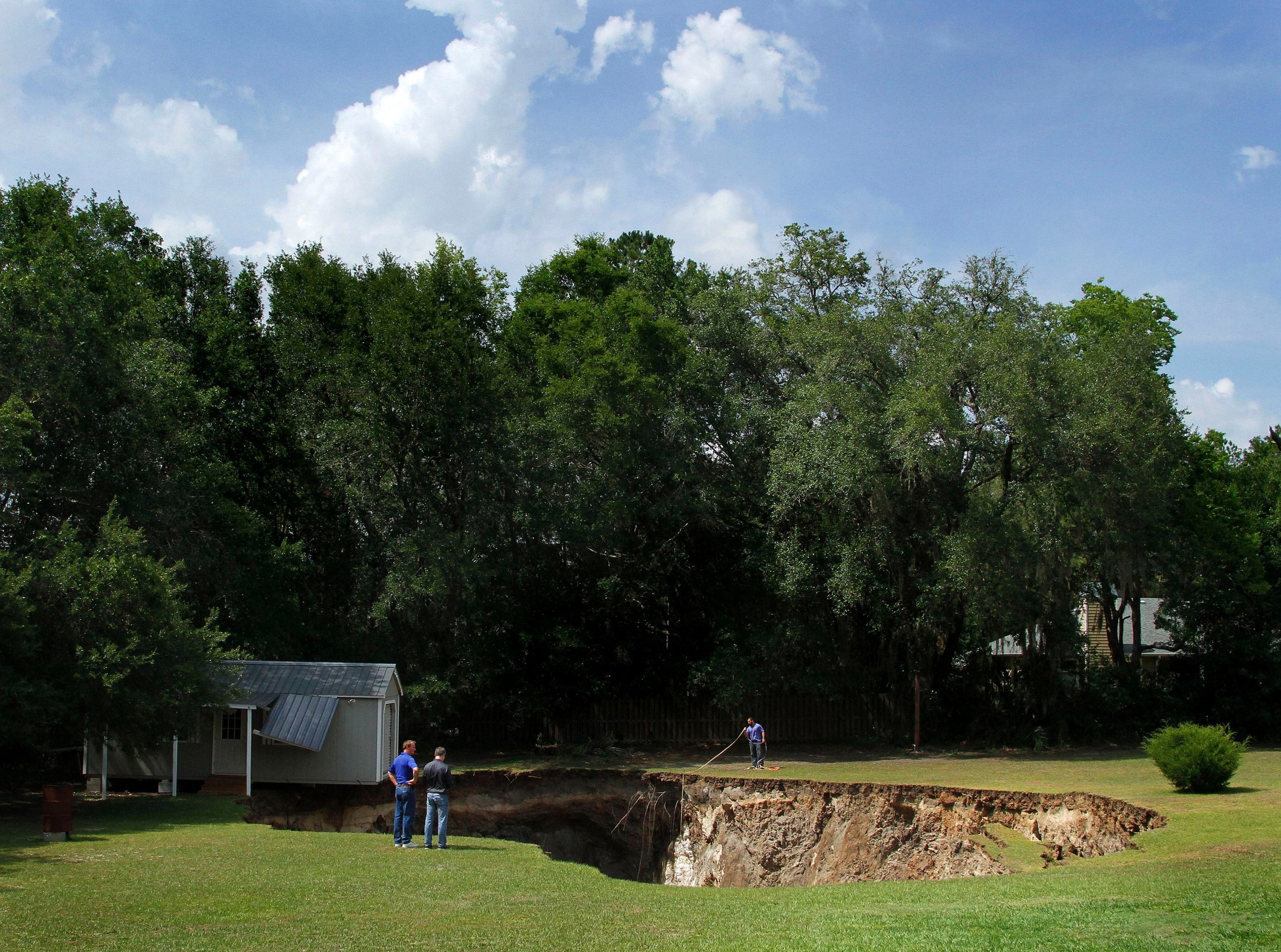 A backyard with a large hole in the ground, three people standing nearby, one person working with a rake, and a small white shed with a metal roof, surrounded by green trees under a partly cloudy sky.