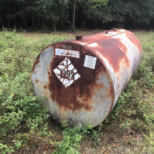 Old, rusted metal fuel tank lying on grass near trees.