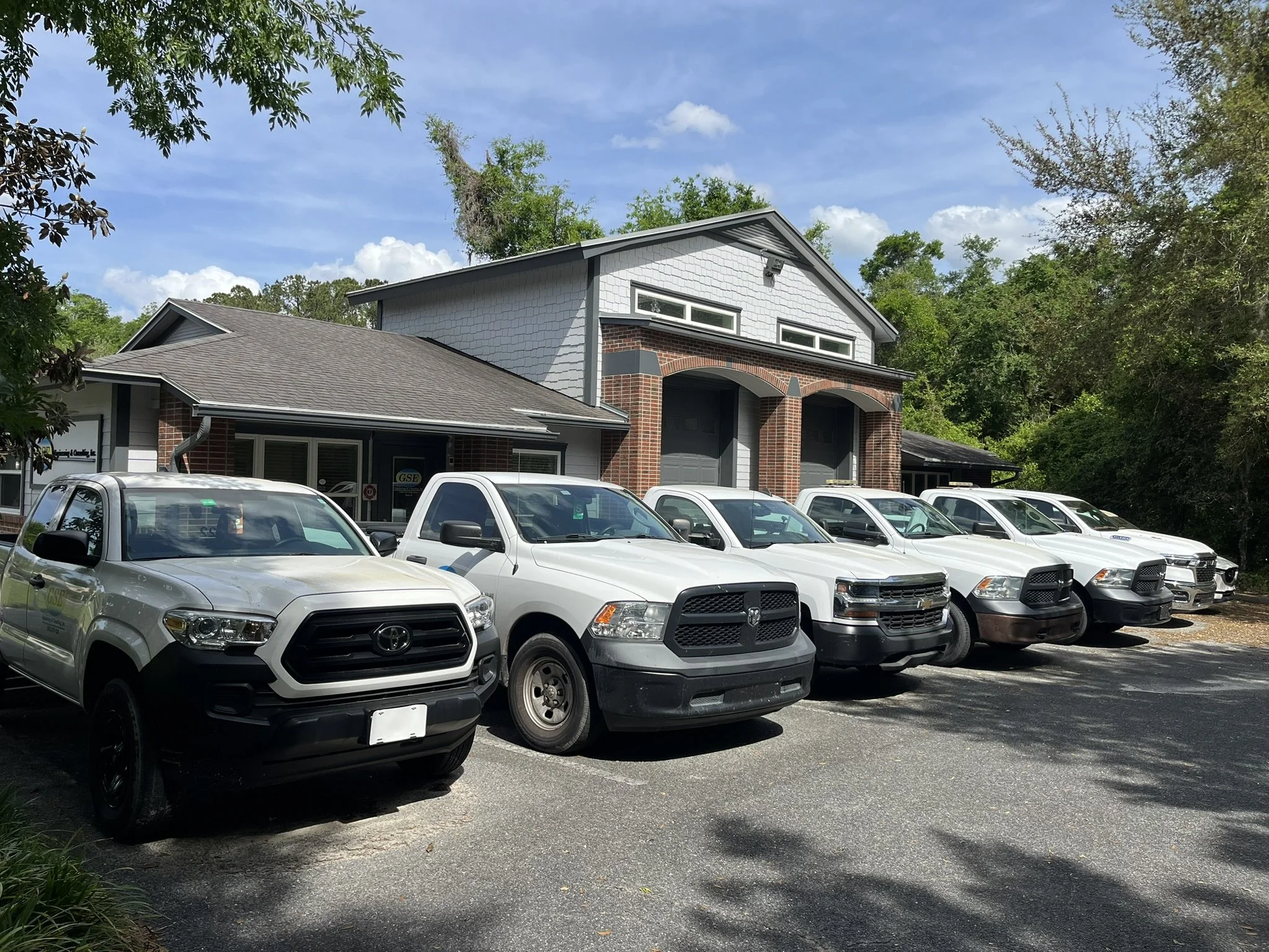 A row of white pickup trucks parked in front of a modern house with brick and white siding exterior, surrounded by trees and greenery, under a partly cloudy sky.