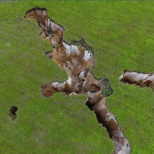 A large hole or sinkhole in a grassy field with exposed soil and underground features.