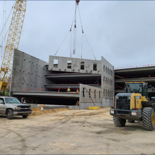 Construction site with a building under construction, crane lifting concrete panels, and construction vehicles like a pickup and a skid-steer loader.