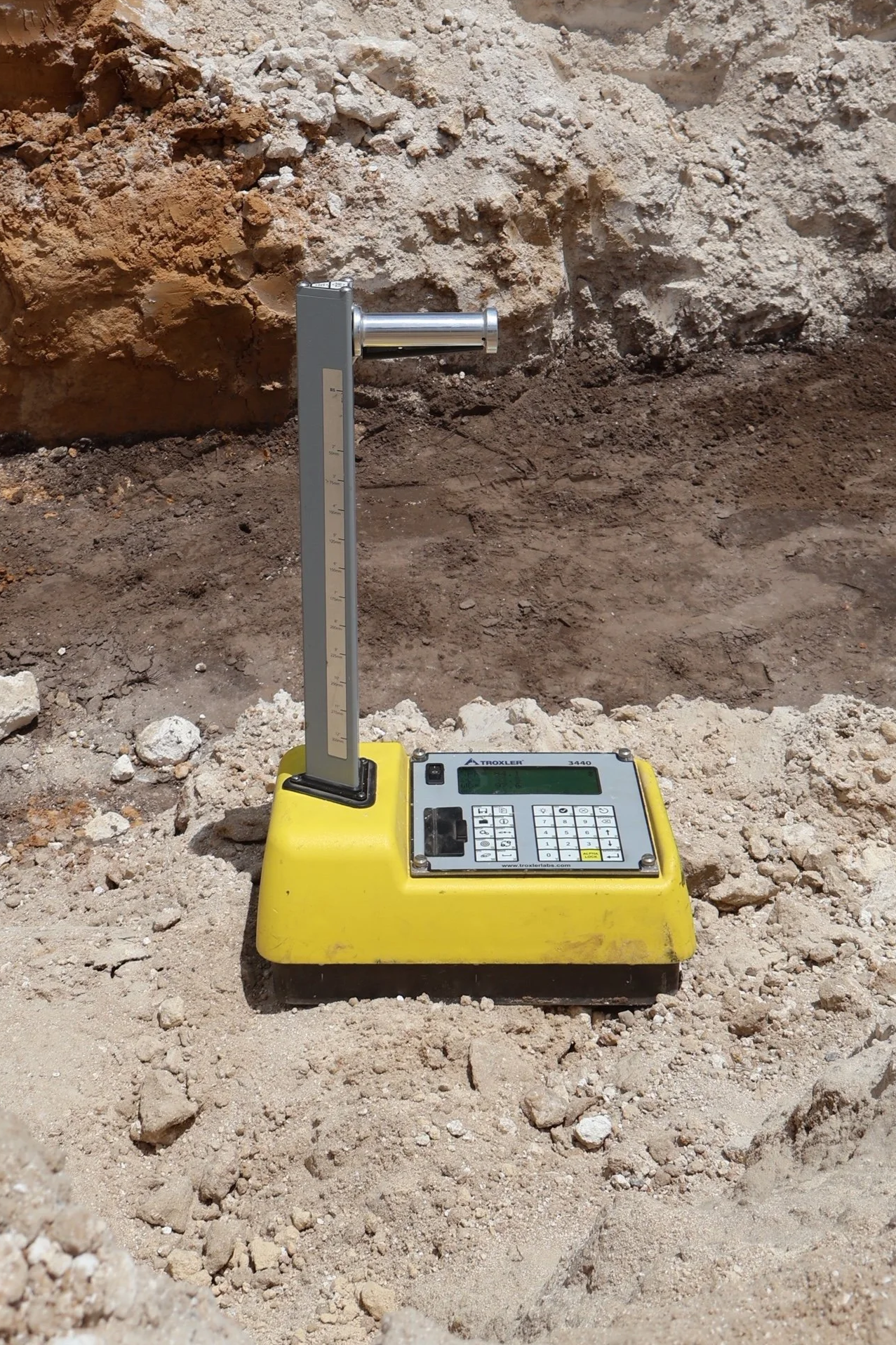 A digital level on yellow base measuring horizontal slope in a rocky construction site, with a background of dirt and soil layers.
