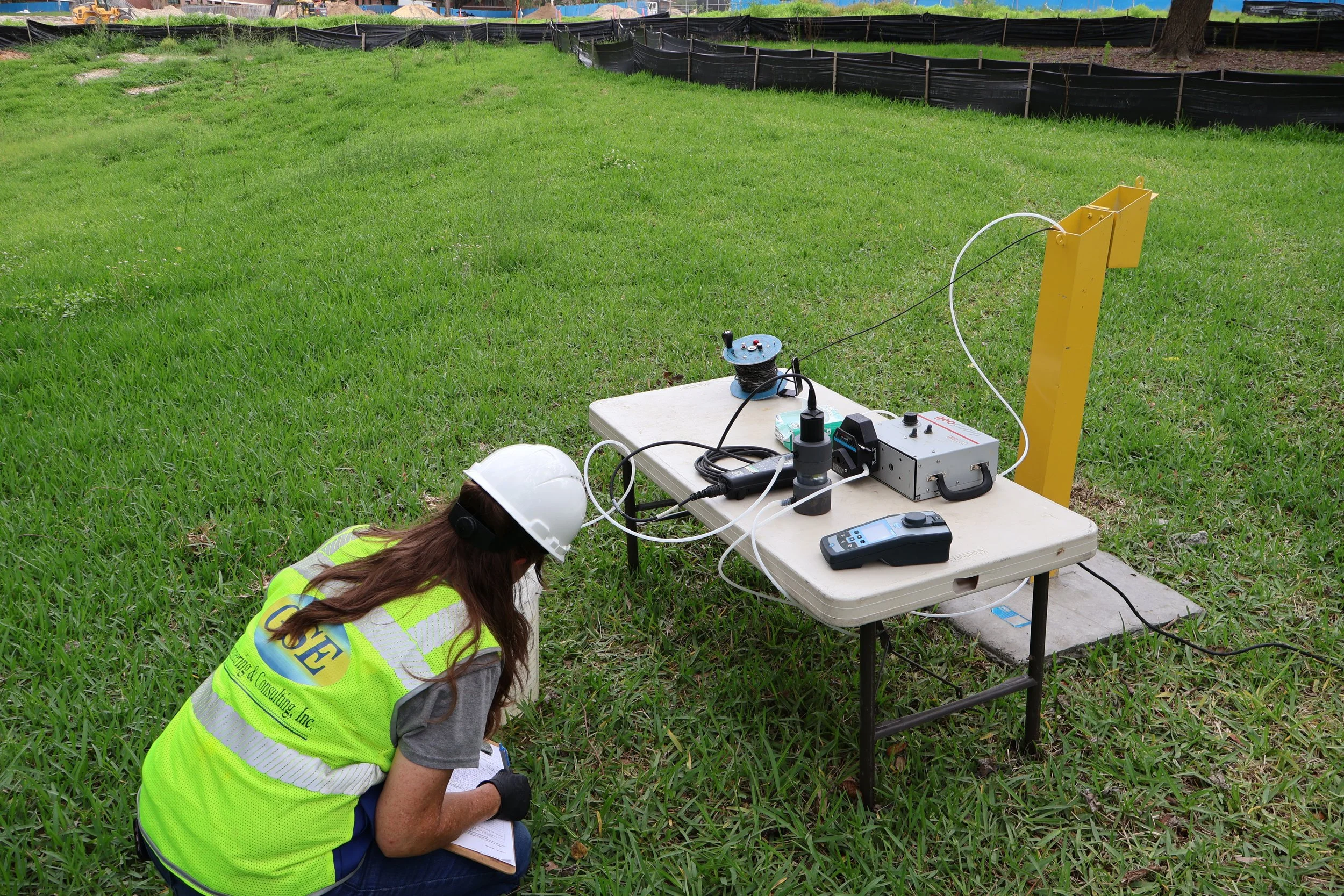 A person wearing a yellow safety vest and white hard hat is crouching on the grass, taking notes on a clipboard. Nearby, there is a table with electronic equipment and tools, including a spool of cable, a remote control, and other devices. In the background, blue sky and trees are visible, along with a black barrier.