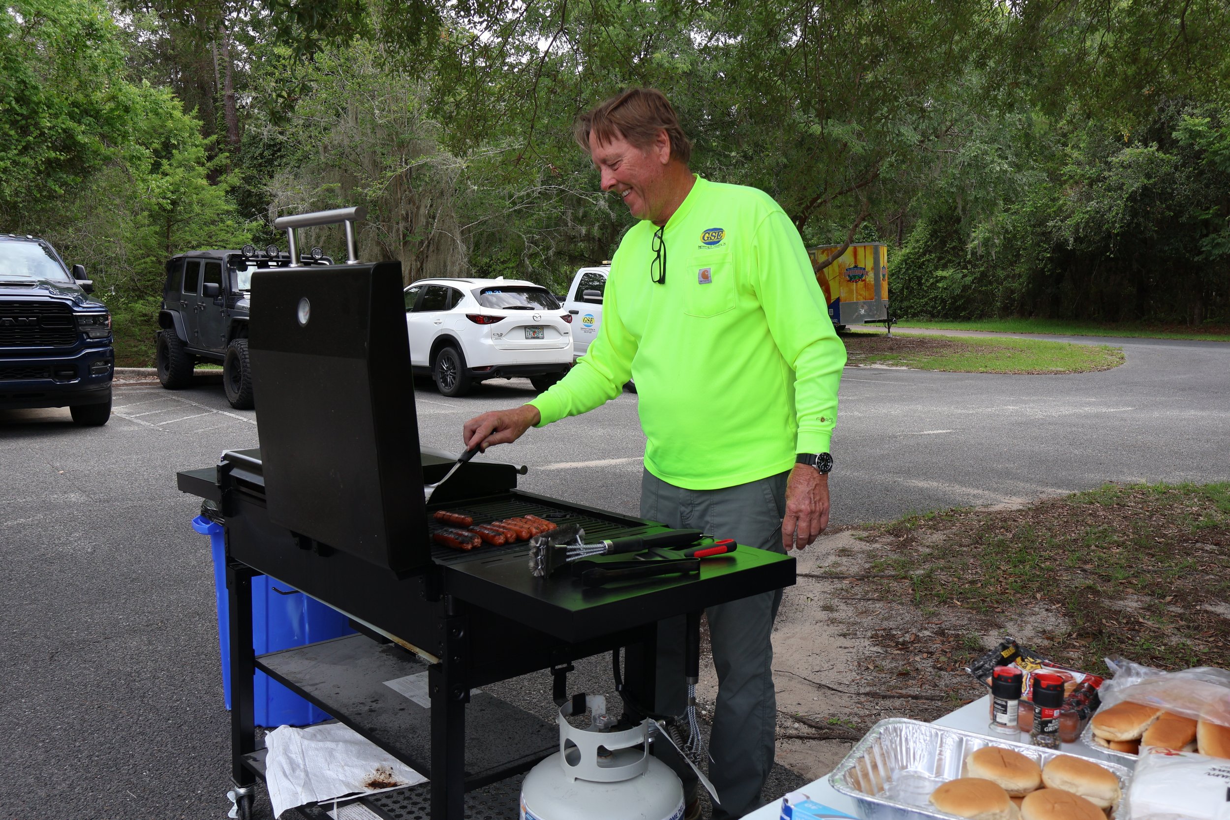 A man in a bright yellow shirt is grilling hot dogs on a barbecue in a parking lot surrounded by trees, with several parked vehicles and a food trailer in the background.
