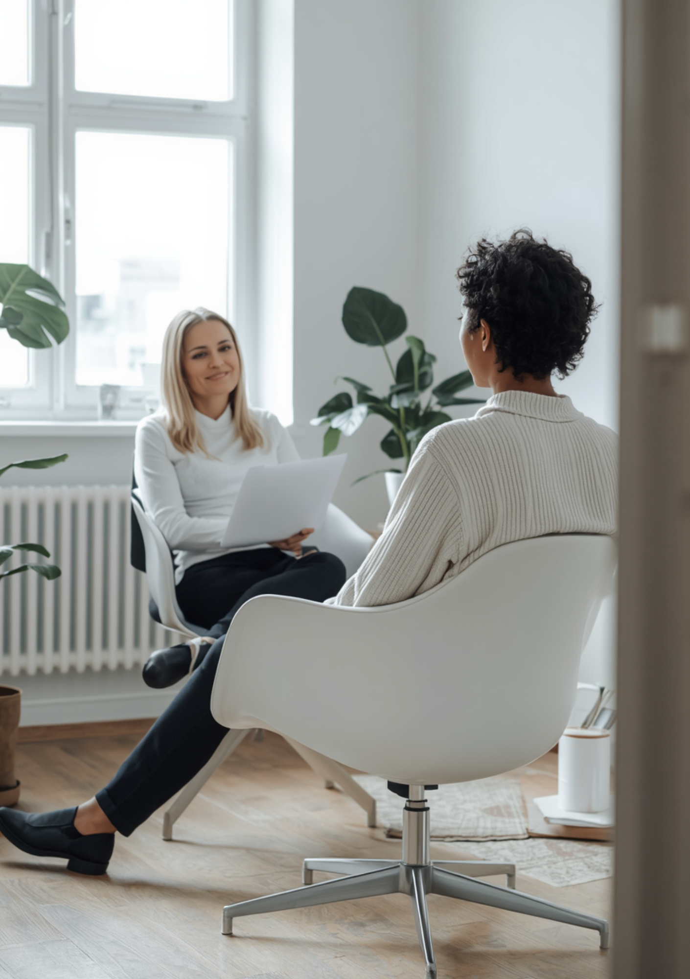 Two women having a conversation in a modern office with large windows and indoor plants.