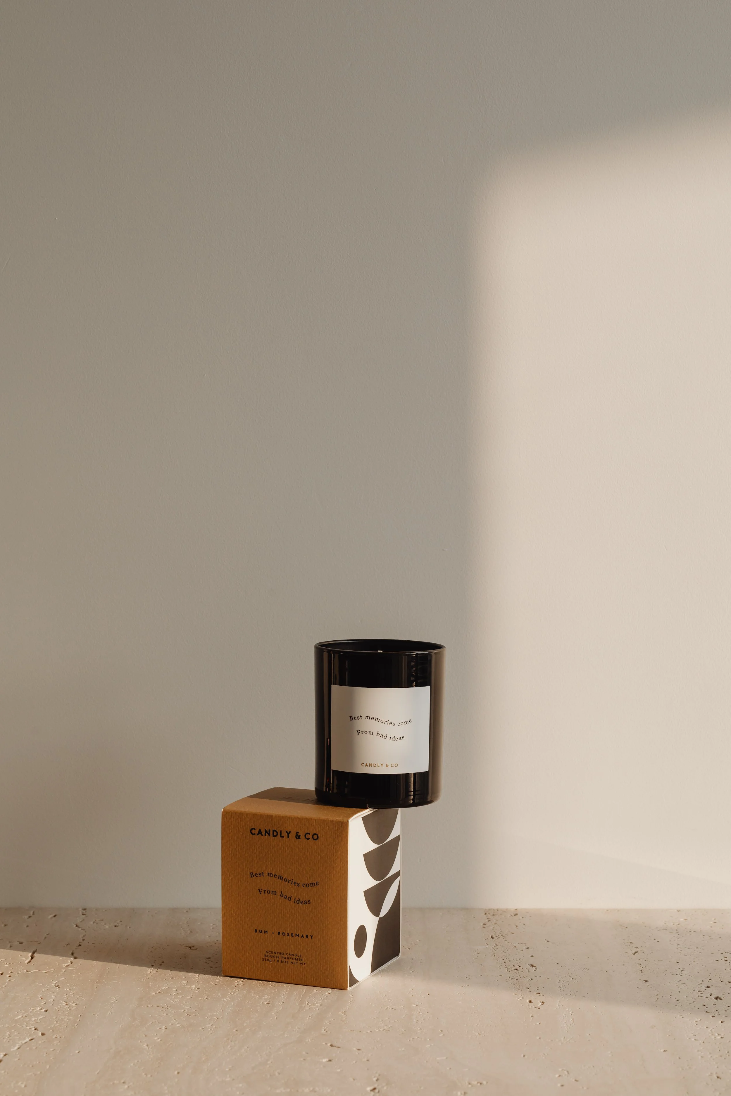 A black scented candle in a glass container with a white label, placed on top of a cardboard box with an abstract design, on a light-colored textured surface against a plain light wall.