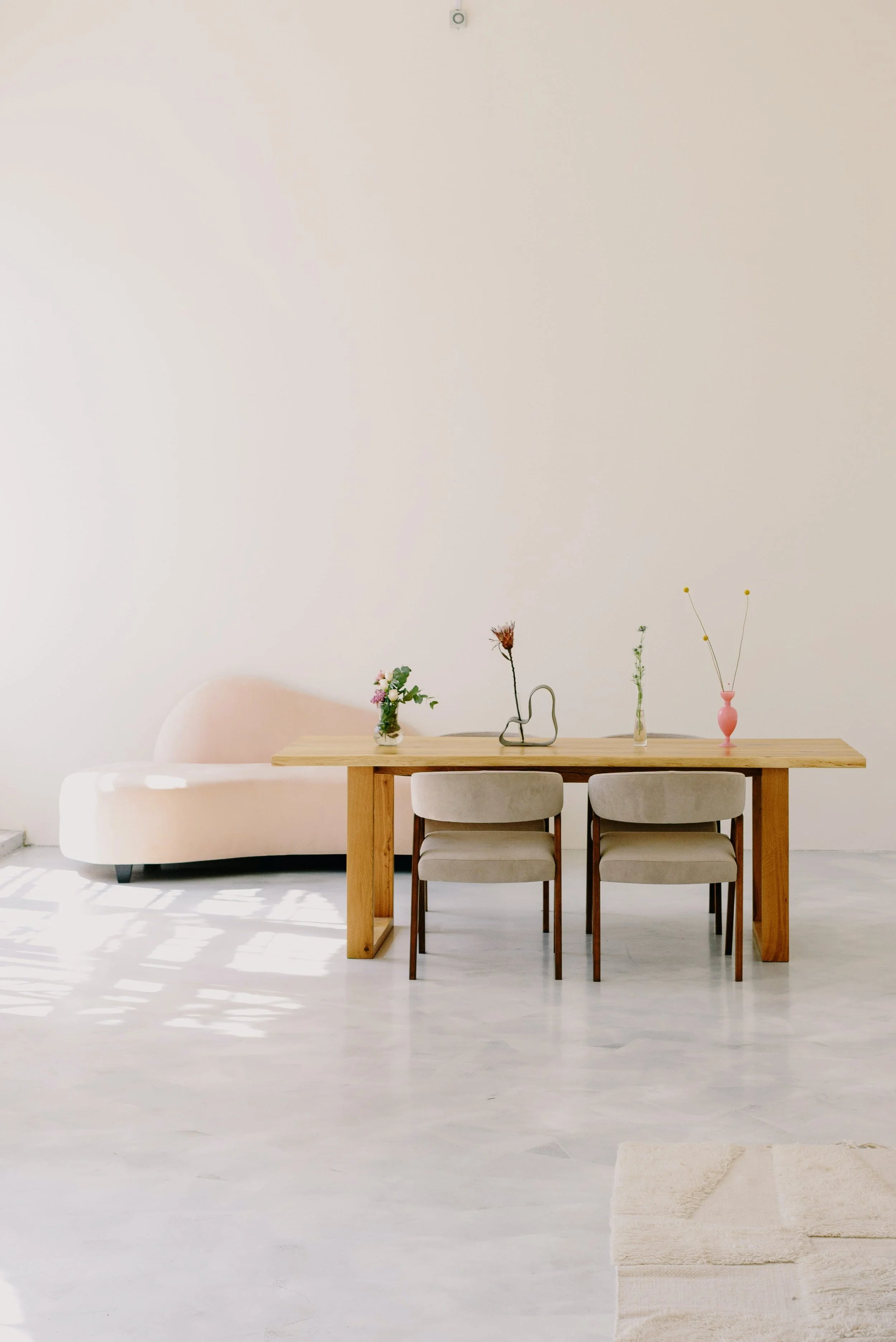 Minimalistic room with a wooden dining table with three chairs, a beige sofa, a white rug, and four vases with flowers on the table, in front of a plain off-white wall.
