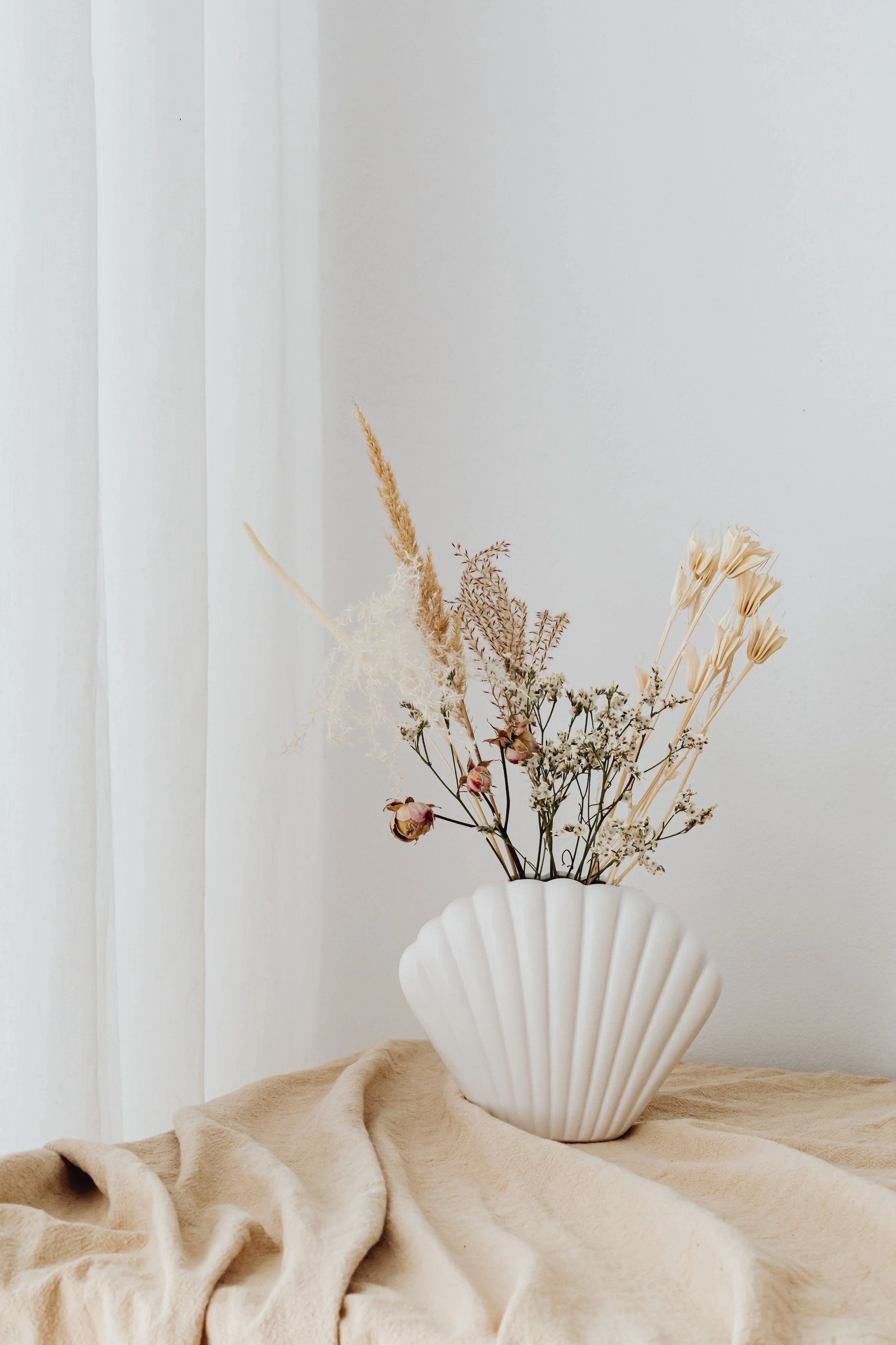 A white ceramic vase with a scalloped design containing dried flowers, placed on a beige fabric surface near a white wall and curtain.