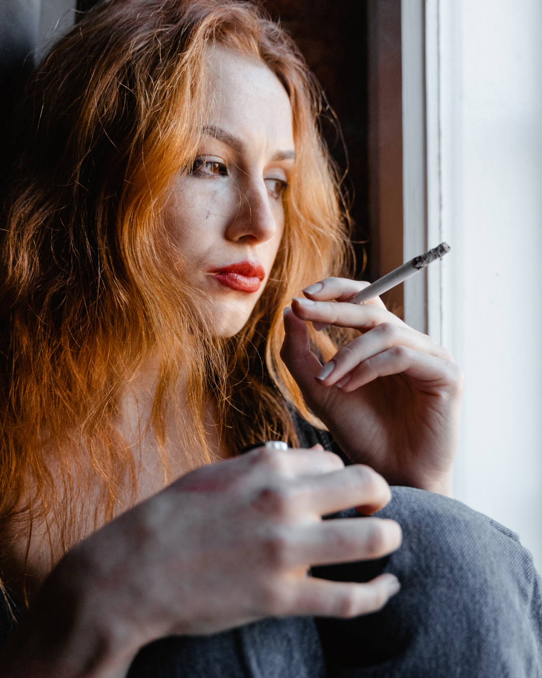 A young woman with long red hair, wearing makeup, holding a cigarette near her mouth, and gazing out of a window.