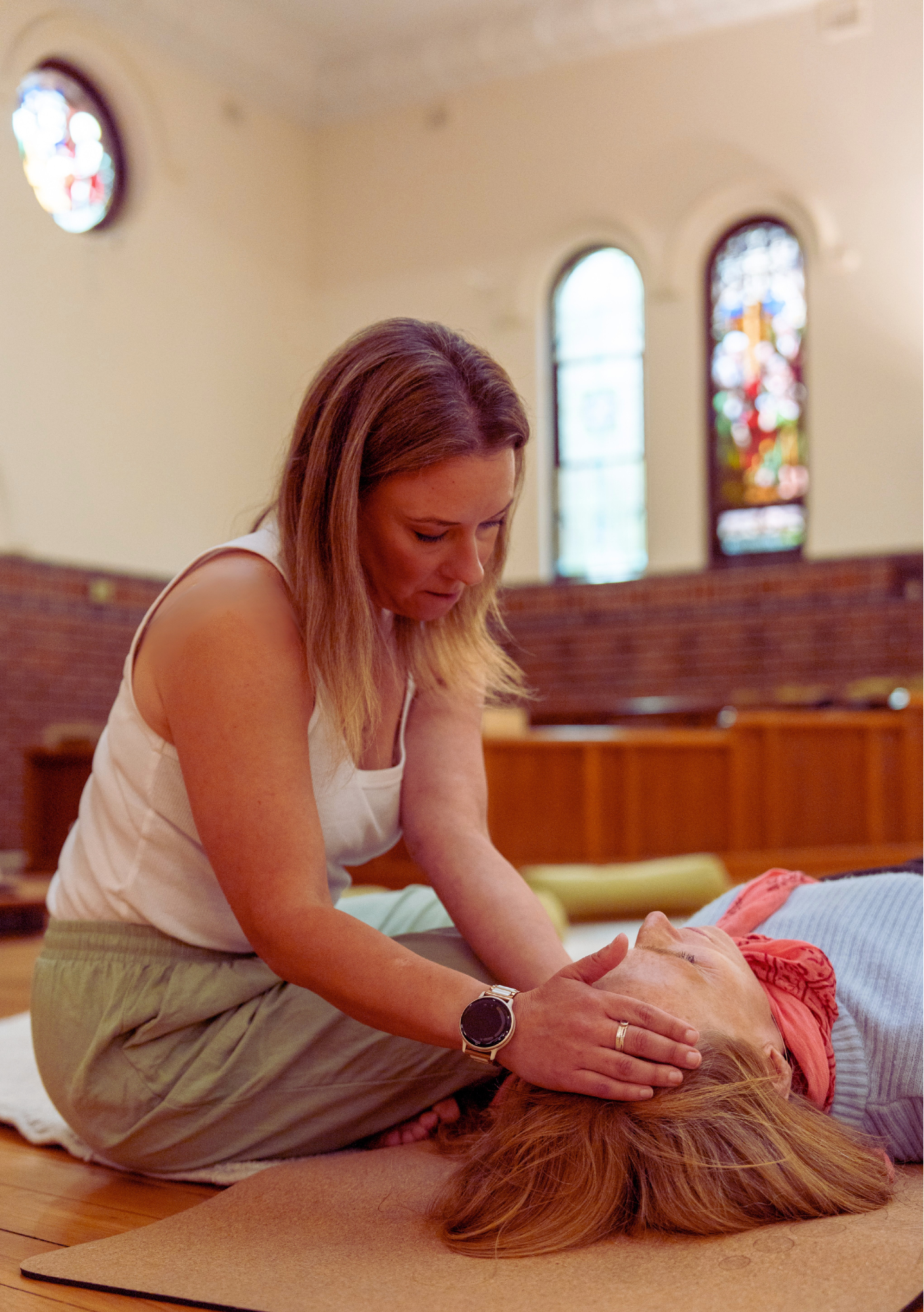 A woman performing facilitated breathwork on a woman lying on a yoga mat in a church setting with stained glass windows in the background.