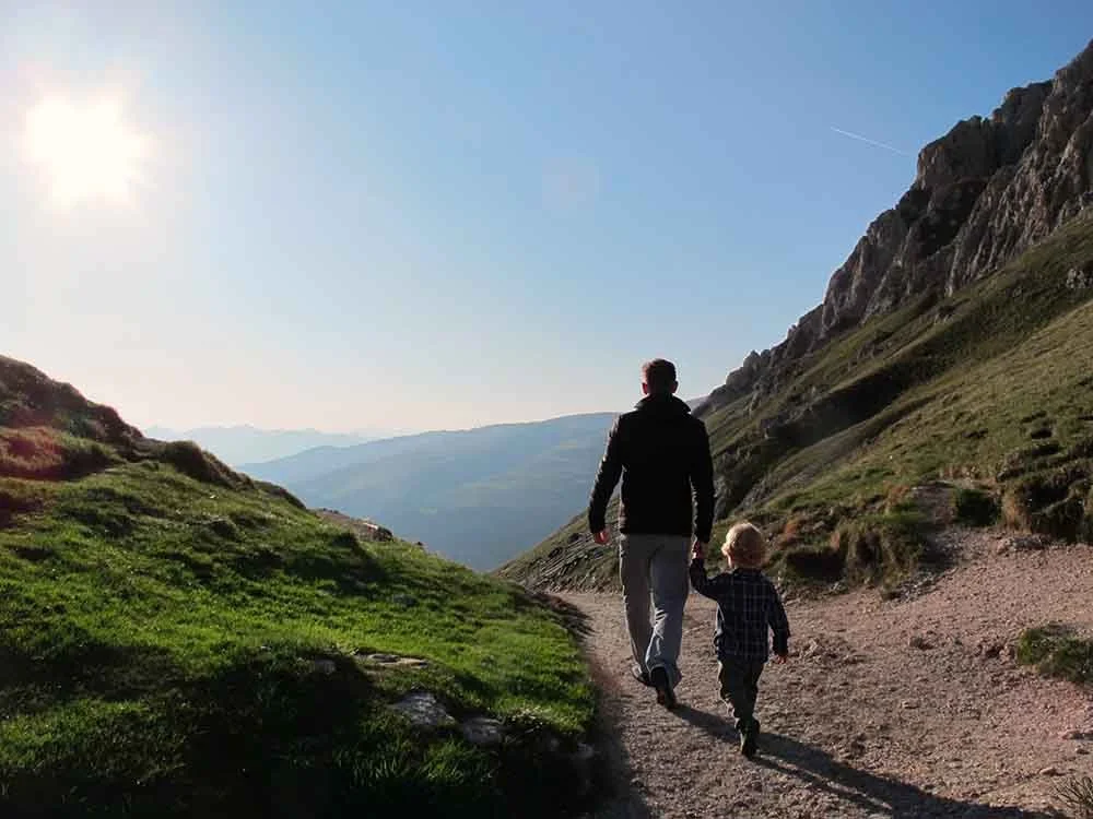 A man and a young boy walking hand in hand on a trail through a mountainous landscape in the late afternoon or early evening, with the sun low in the sky.