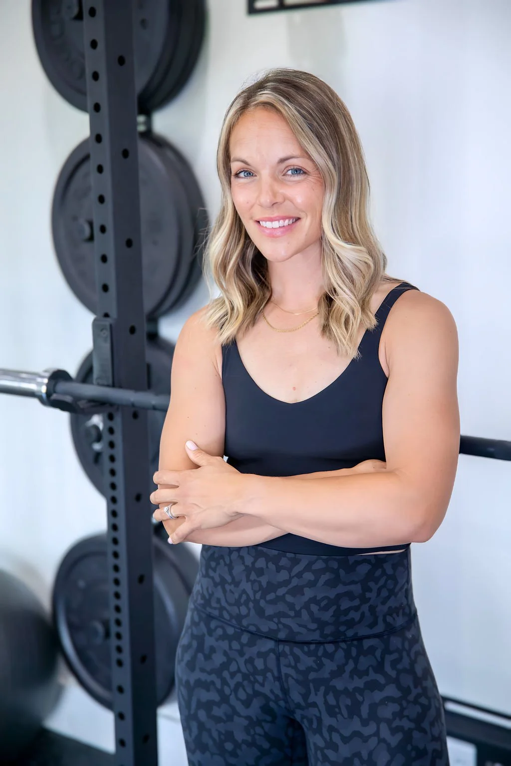 A smiling woman with blonde wavy hair standing in a gym near weightlifting equipment, wearing a black sports top and patterned workout pants.