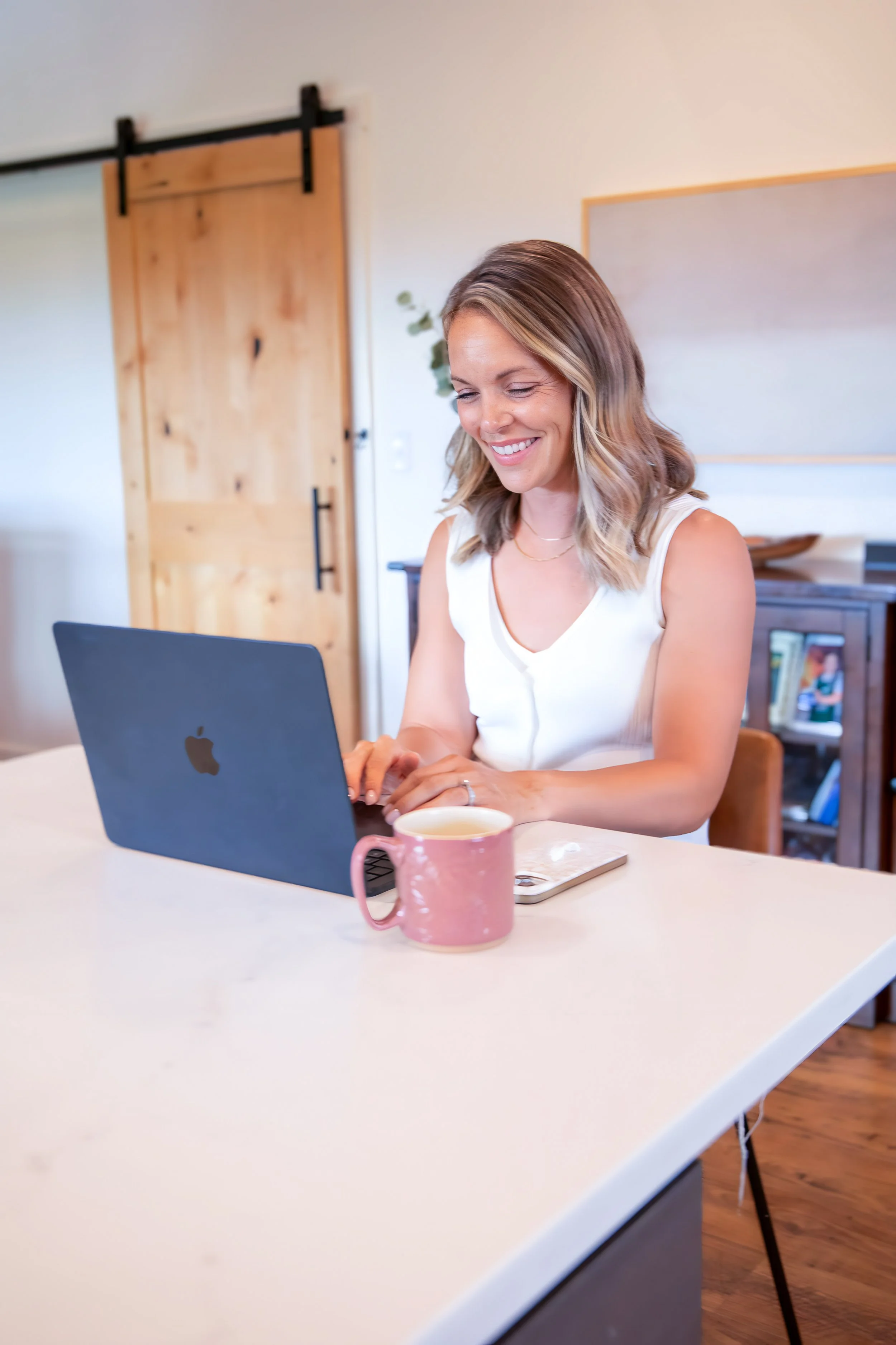 A woman sitting at a white table working on a laptop, with a pink mug in front of her, in a cozy room with a barn door and bookshelf in the background.