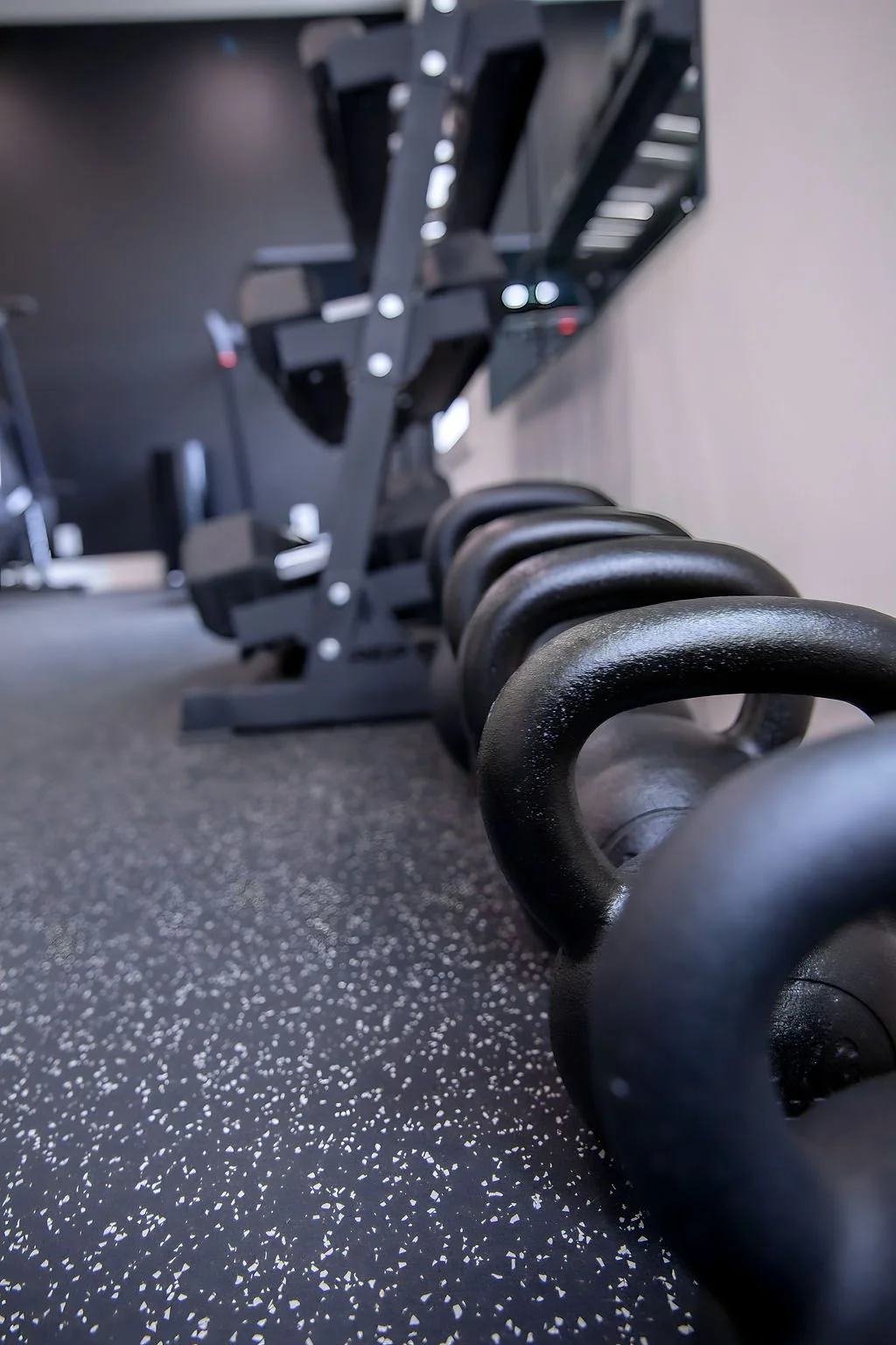 Close-up view of a row of black kettlebells on the floor at a gym, with exercise equipment and monitors in the background.