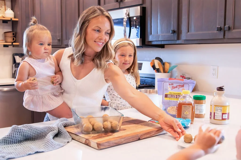 A woman and two young girls in a kitchen preparing food. The woman is holding a girl and reaching for food on the counter, with baking ingredients and utensils around.