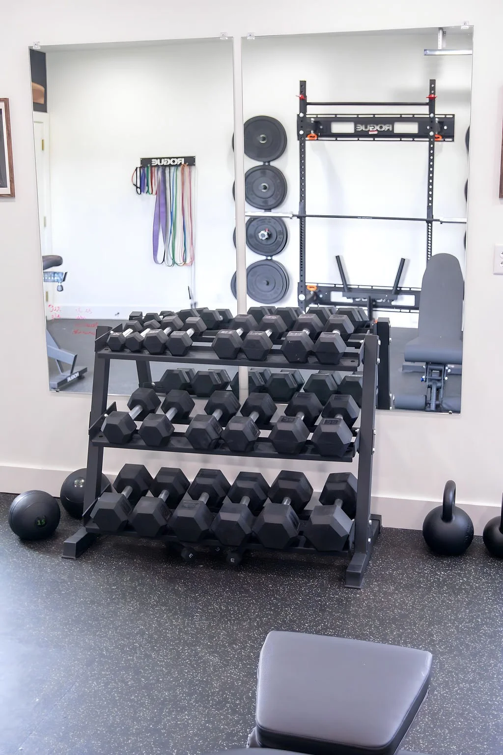 Gym weight room with a rack of black dumbbells, medicine balls on the floor, a mirror reflecting resistance bands, weight plates, and a wall-mounted pull-up bar.