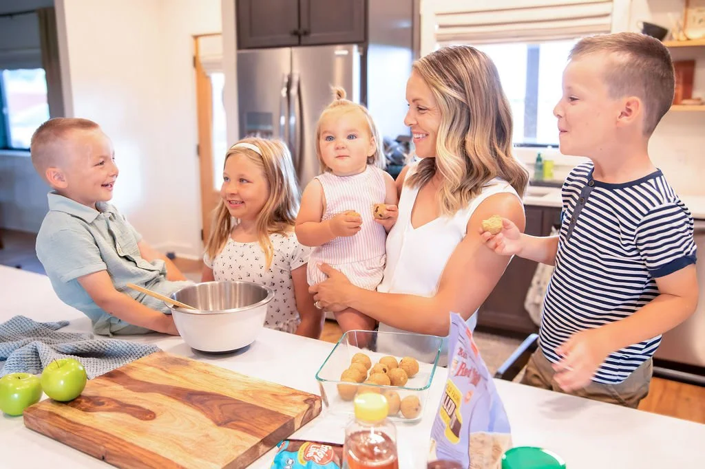 A woman and four children in a kitchen preparing cookies, with baking ingredients and apples on the counter.