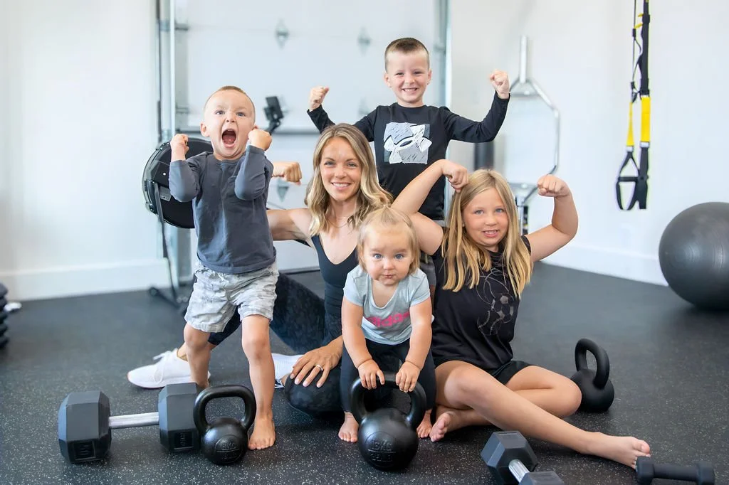 A woman with four children in a gym, all showing off strength by flexing muscles or posing with fitness equipment such as kettlebells and dumbbells, with fitness gear and equipment in the background.