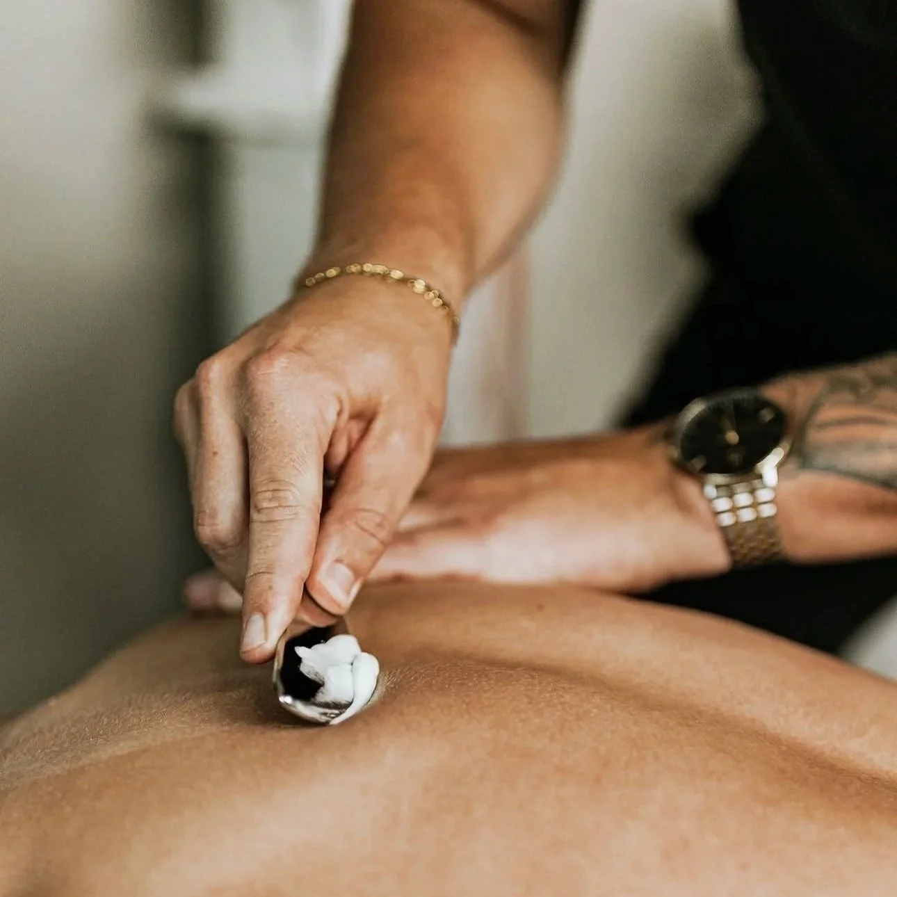 A person is giving a massage with cream on a client's back. The person has a bracelet and a watch, and is using a small spoon to apply cream.