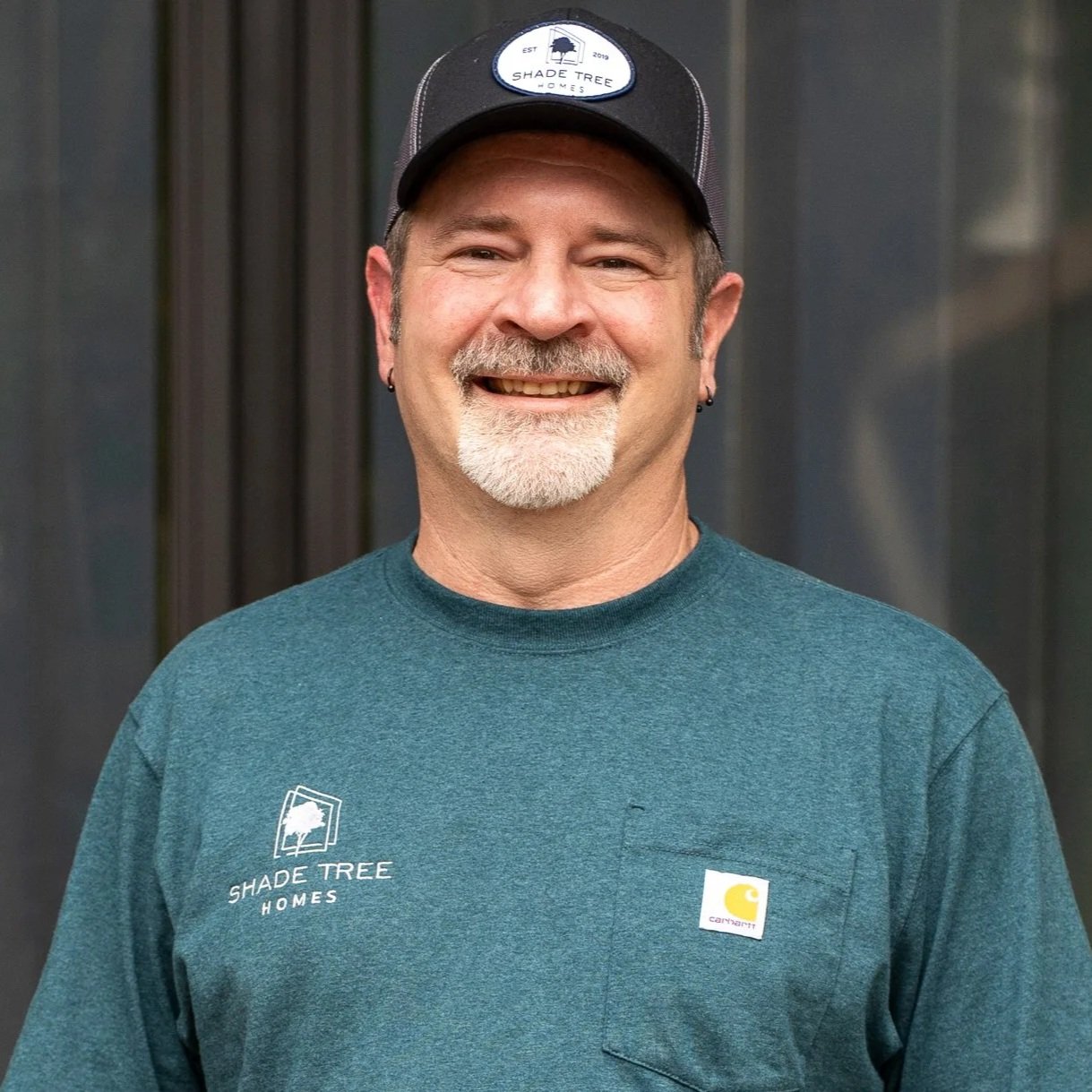 A man smiling wears a black cap with the Shade Tree Homes logo and a teal Carhartt shirt with the same logo on the chest, standing in front of a dark background.