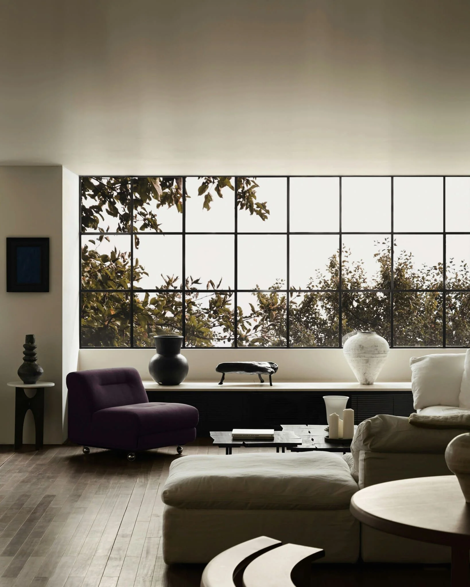Modern living room with large windows, a purple armchair, beige sofas, decorative vases, a side table, and a coffee table, with a view of trees outside.