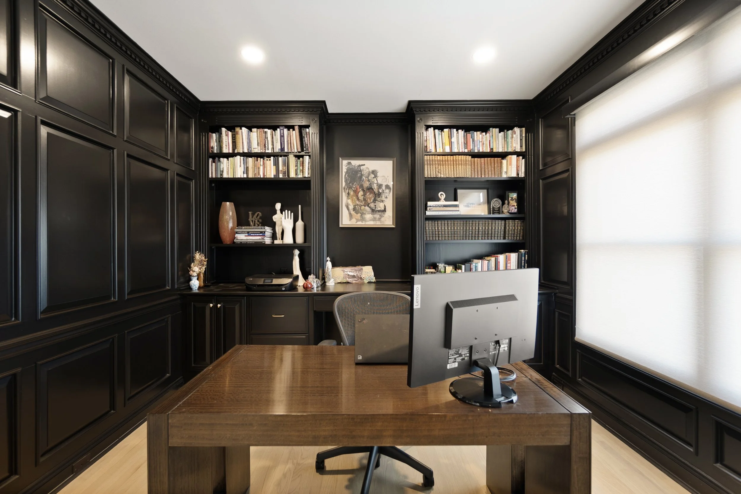 A home office with black paneled walls, built-in bookshelves filled with books, a wooden desk with a computer monitor, and decorative items on the shelves, illuminated by ceiling spotlights and a window with a white curtain.