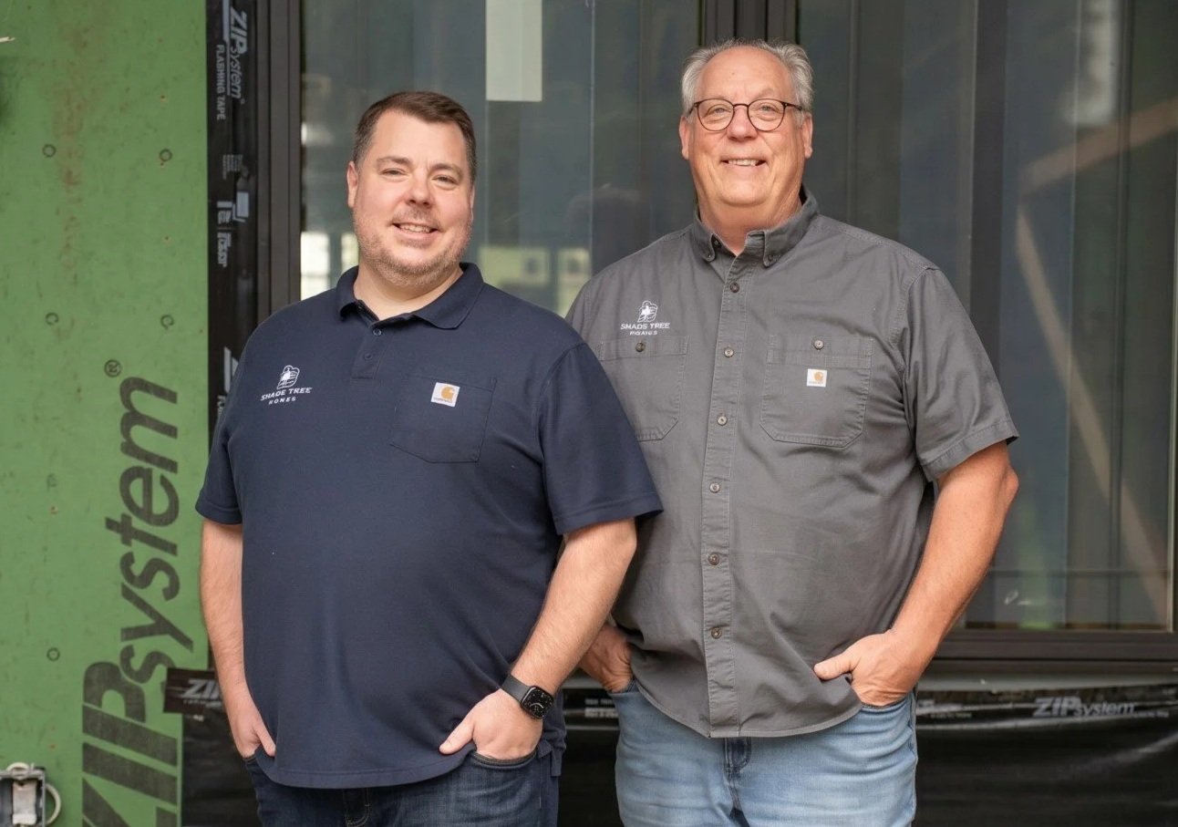 Two smiling men standing in front of a building under construction, wearing work shirts with logos.